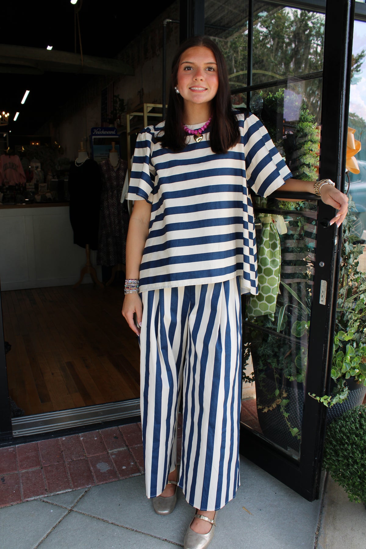 A woman stands in front of a storefront doorway wearing a matching navy and white striped two-piece outfit consisting of a short-sleeve top and wide-leg pants.