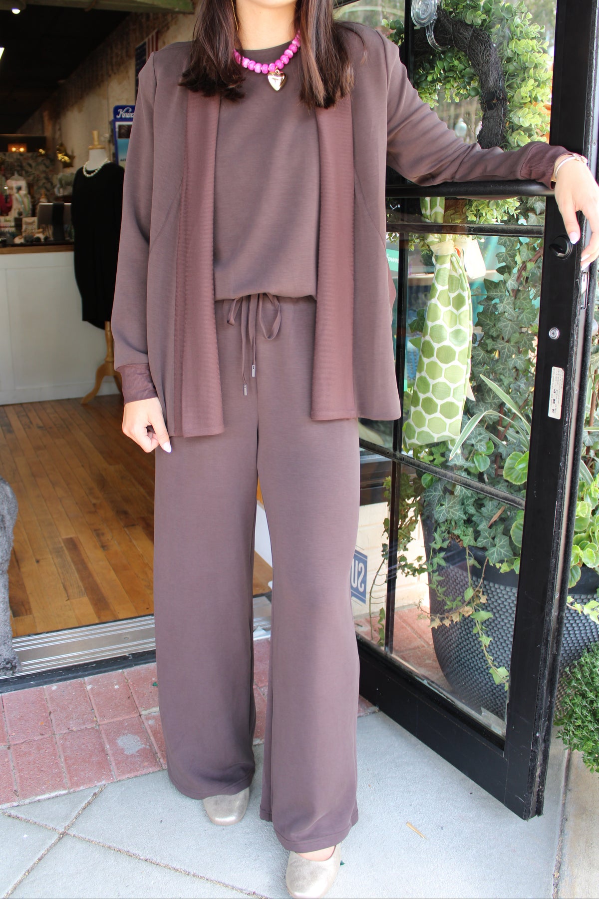 A woman models a matching lounge set consisting of a long-sleeve top, drawstring waist, and wide-leg pants in a dusty mauve-brown color, styled with a matching open-front cardigan and a pink beaded necklace.