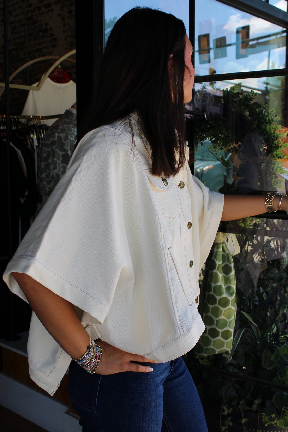 A woman wearing a white short-sleeve button-up shirt with gold buttons, paired with blue jeans, standing in front of a window in a clothing store.