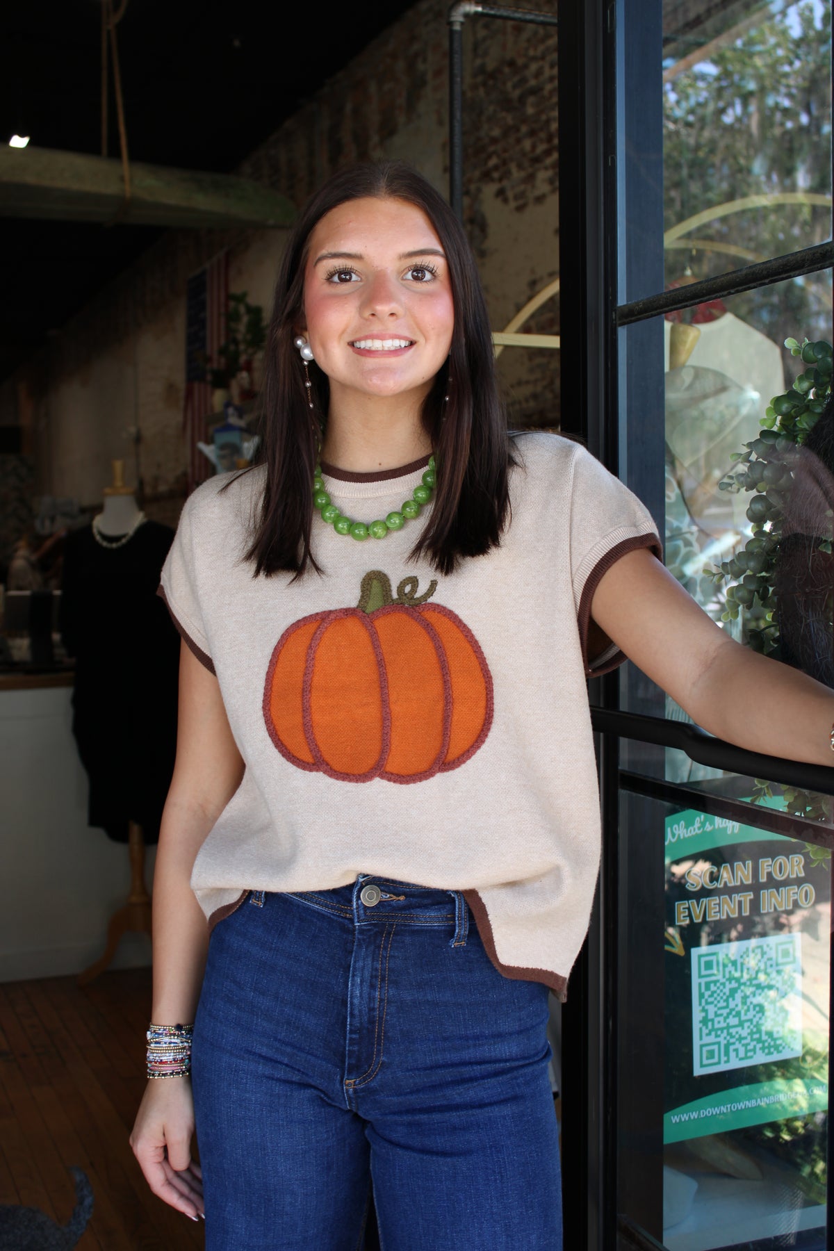 A woman wearing a cream-colored knit sweater with a large orange pumpkin graphic on the front, paired with blue jeans and a green beaded necklace, standing in front of a shop window.