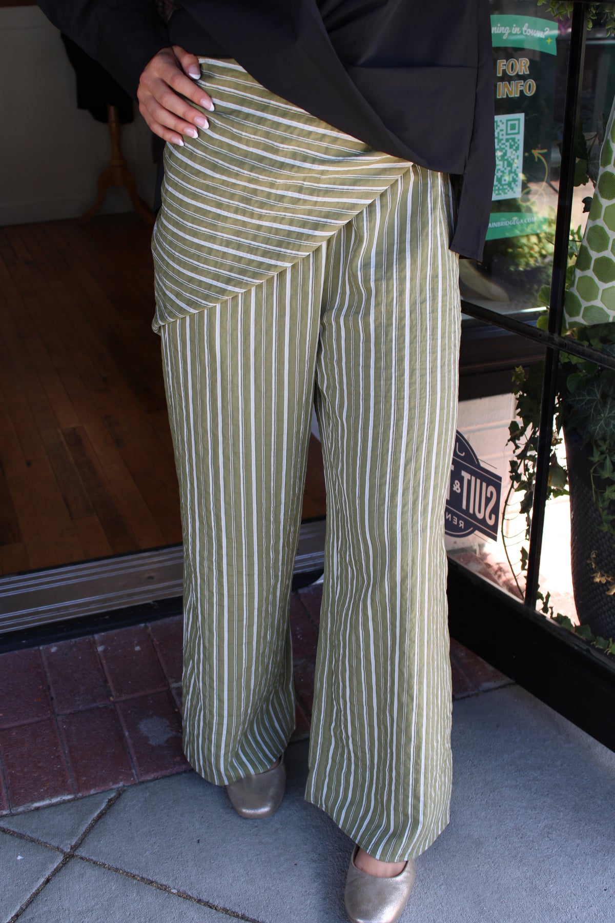 A person wearing wide-leg pinstriped trousers in olive green and white, paired with a black top and metallic silver shoes, standing on a sidewalk.