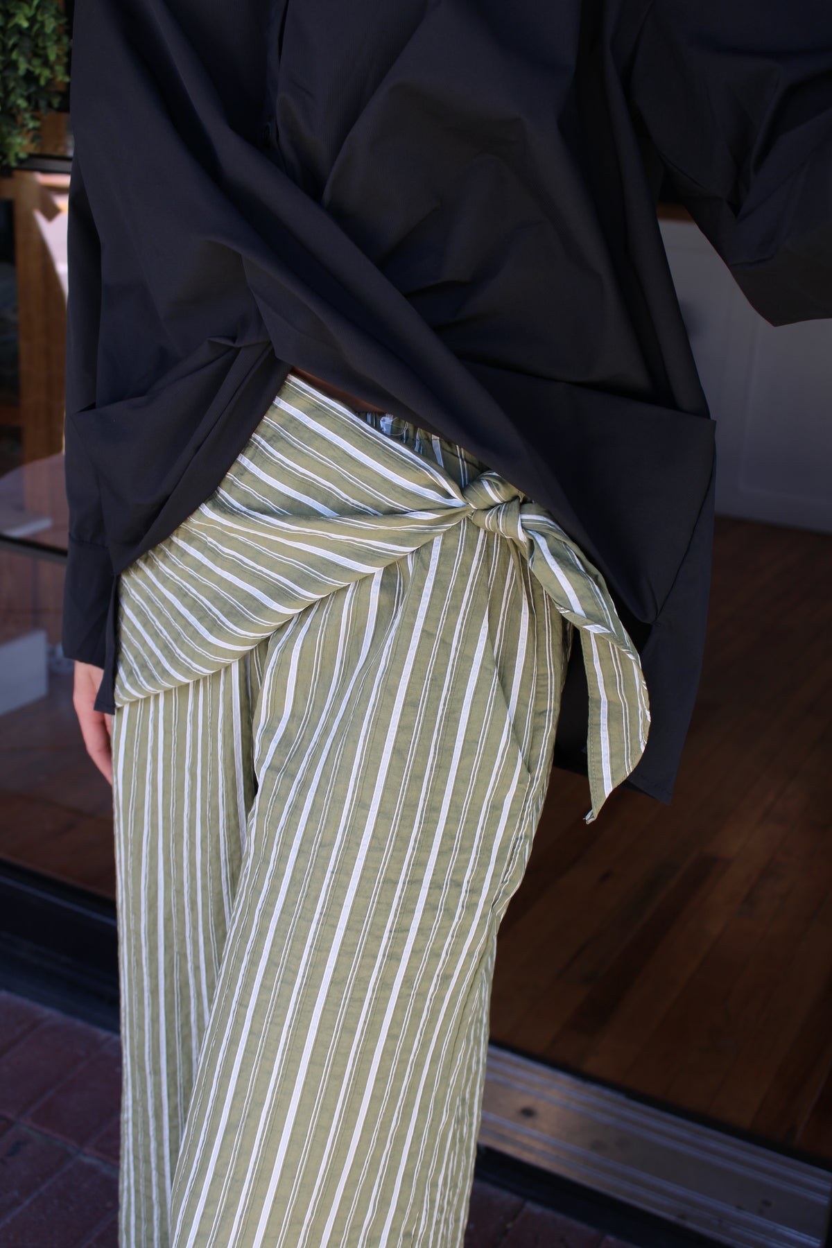 A close-up of a person wearing wide-leg trousers with vertical olive green and white stripes, styled with a black shirt tied around the waist.