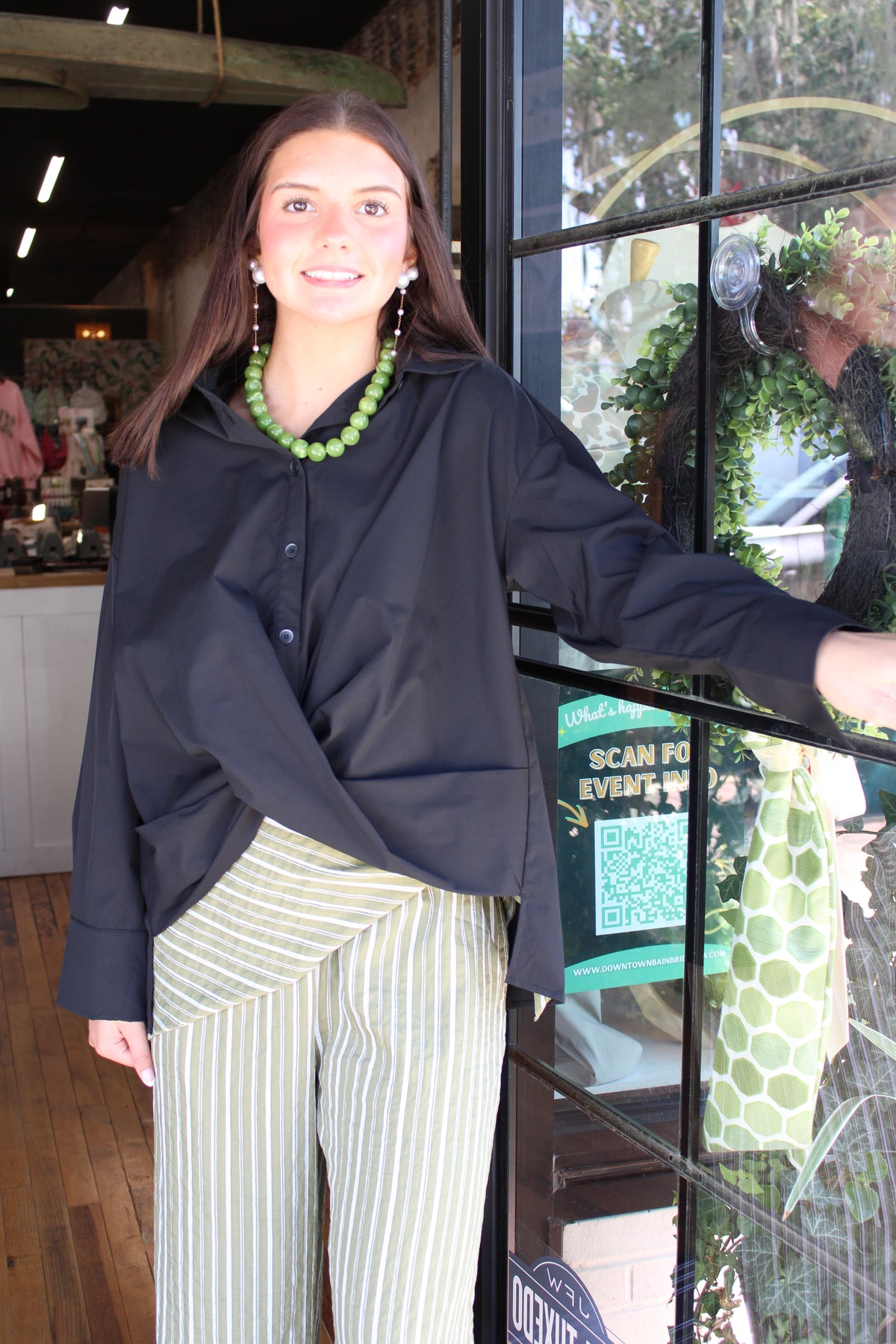 A woman posing in front of a shop window wearing a black long-sleeve button-up shirt and green and white vertically striped pants, accessorized with a chunky green beaded necklace.