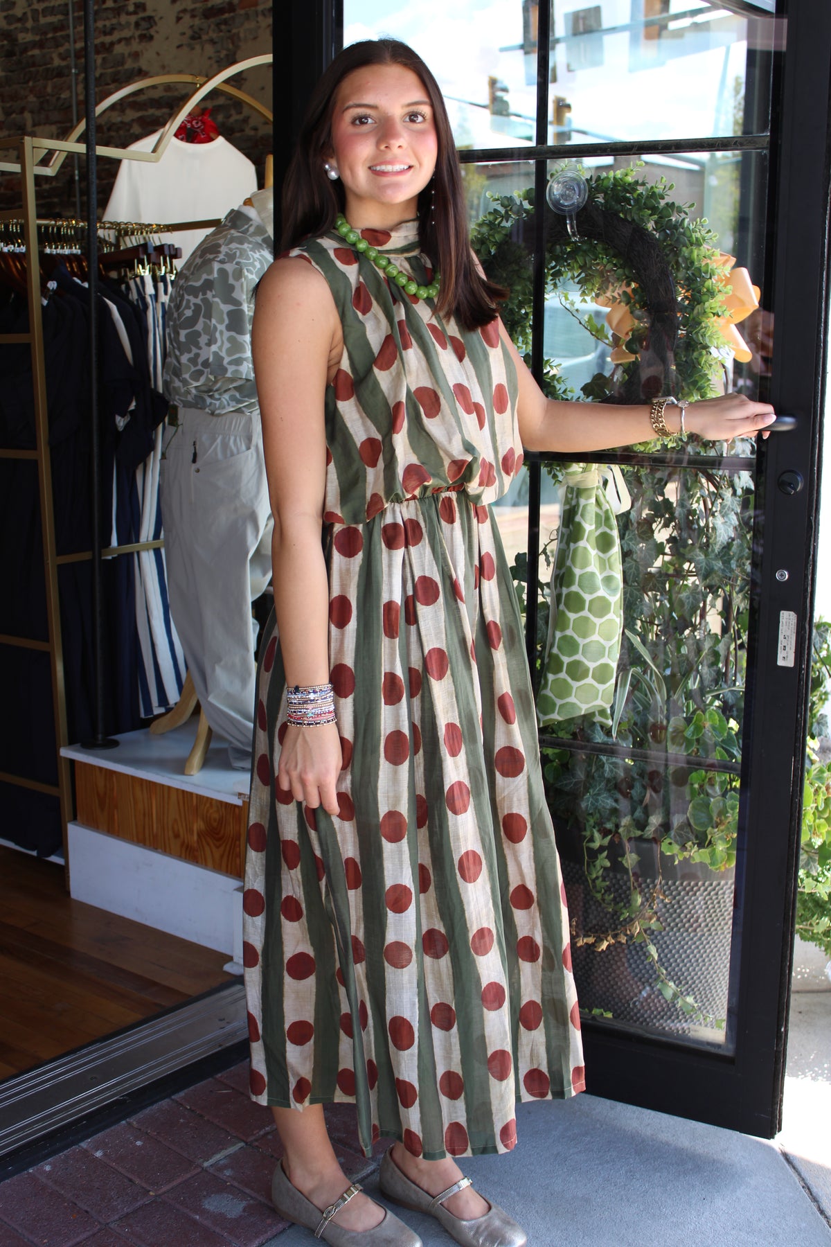 A woman wearing a sleeveless midi dress with vertical olive green and beige stripes and red polka dots, standing in a boutique doorway.