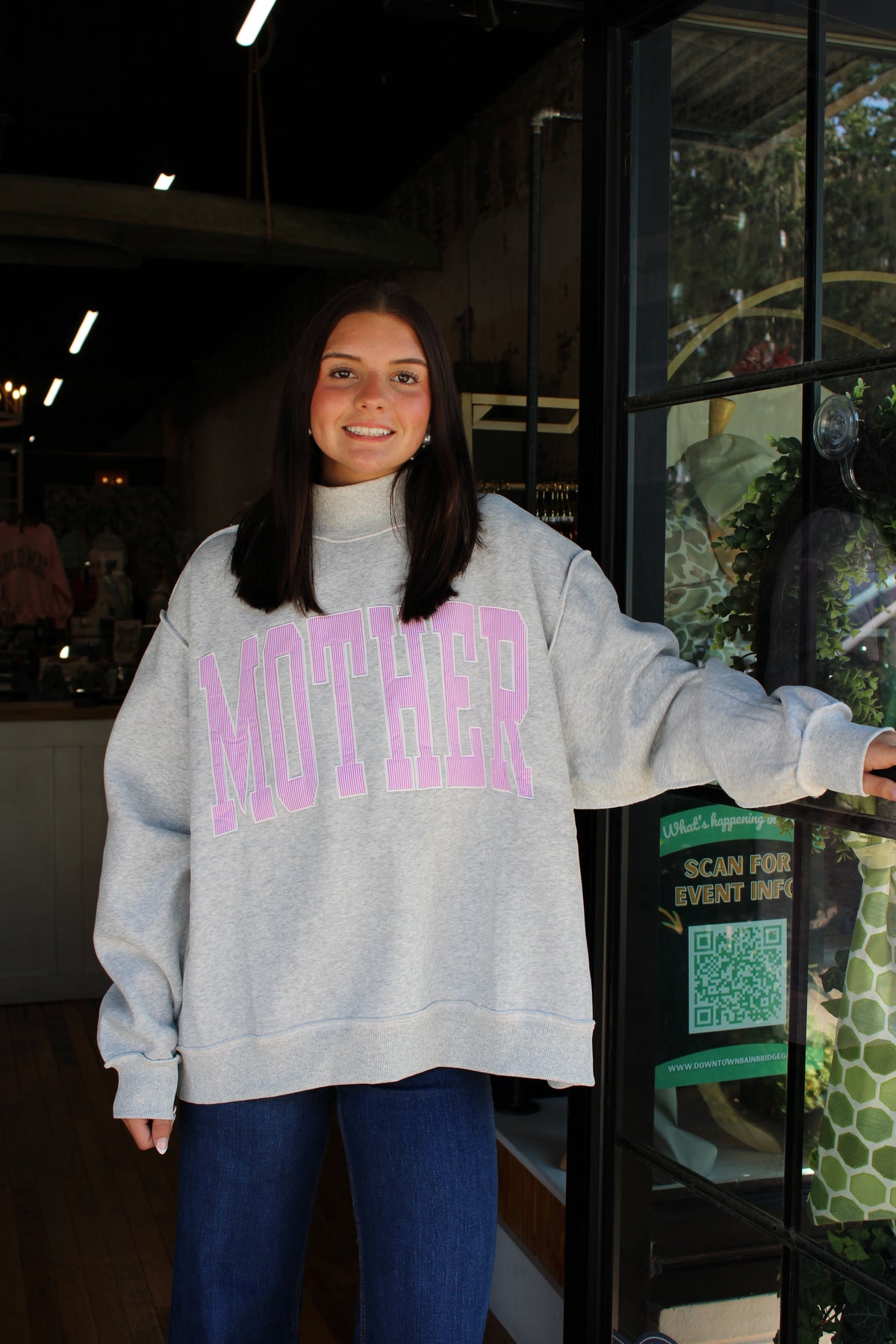 A woman wearing an oversized light gray sweatshirt with 'MOTHER' printed in pink arched letters across the front, standing in front of a glass storefront door.