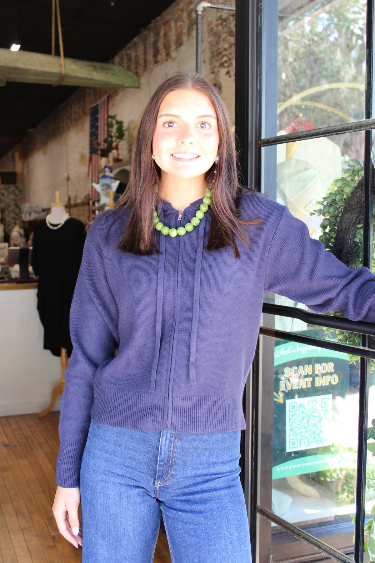 A woman wearing a navy blue knit zip-up hoodie with a drawstring hood and blue jeans, standing in a shop next to a window.