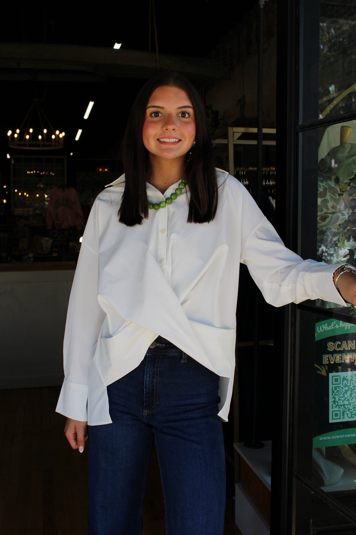 A woman with dark hair stands in a shop doorway wearing a white long-sleeve button-up shirt with a twisted front detail and dark blue jeans.