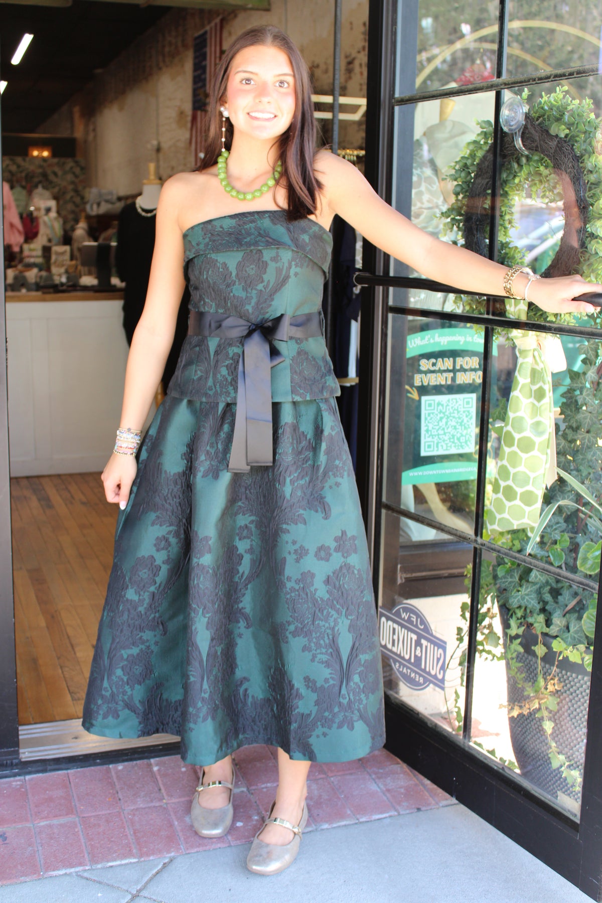 A woman wearing a strapless midi dress with a dark green and black damask pattern, a matching waist sash, and a large bow at the waist, posing in front of a boutique storefront.