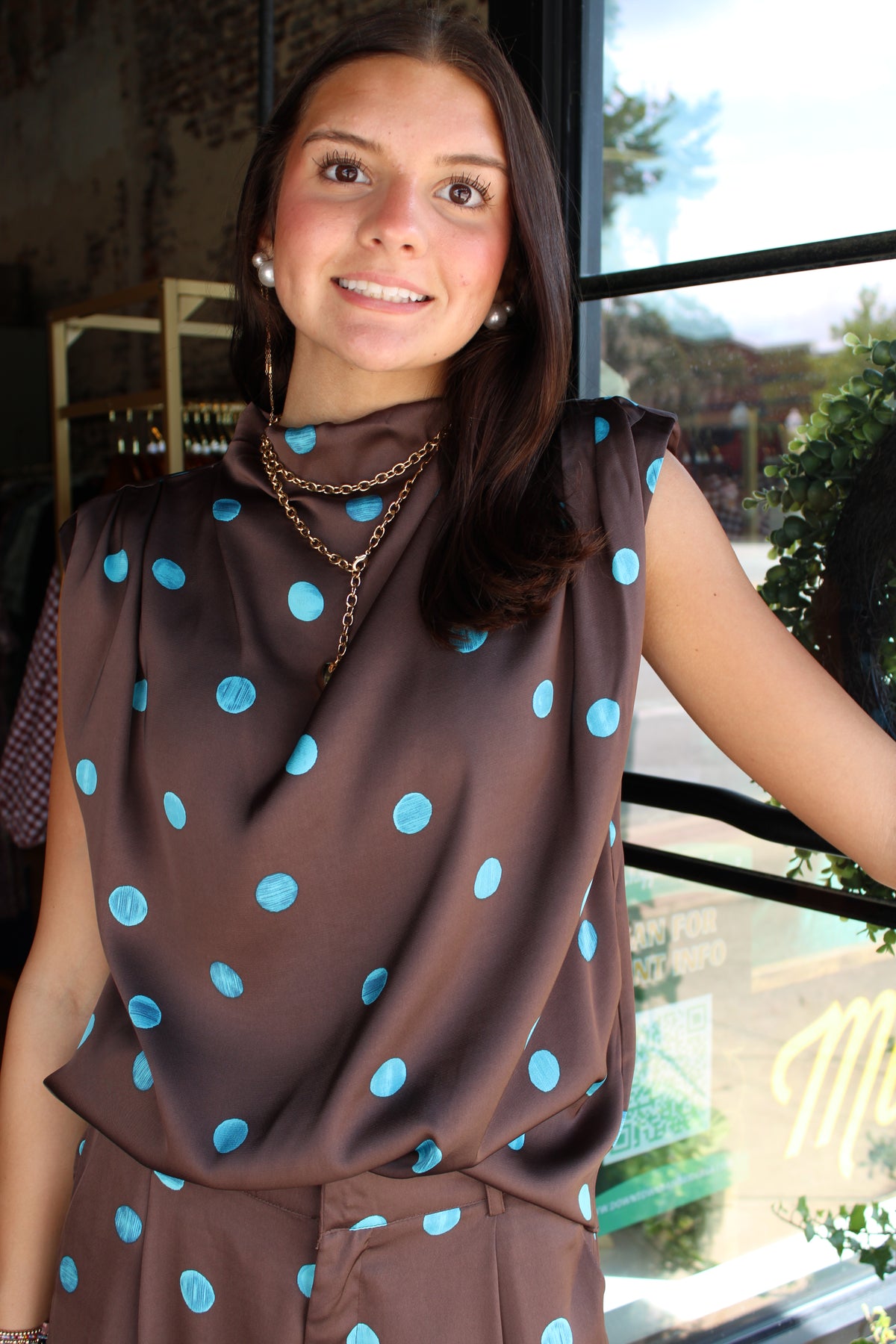 A woman wearing a brown sleeveless top with light blue polka dots, standing in front of a window.
