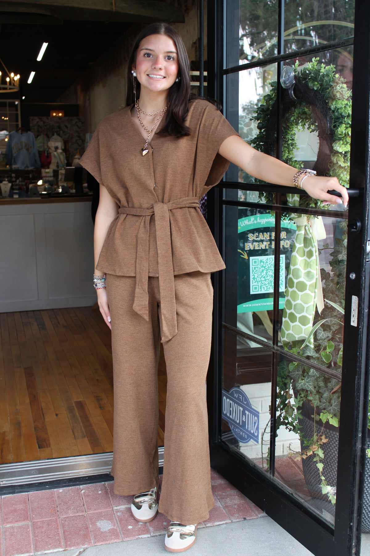 A woman wearing a brown two-piece matching set with a short-sleeve top and wide-leg pants, standing in the doorway of a clothing store.