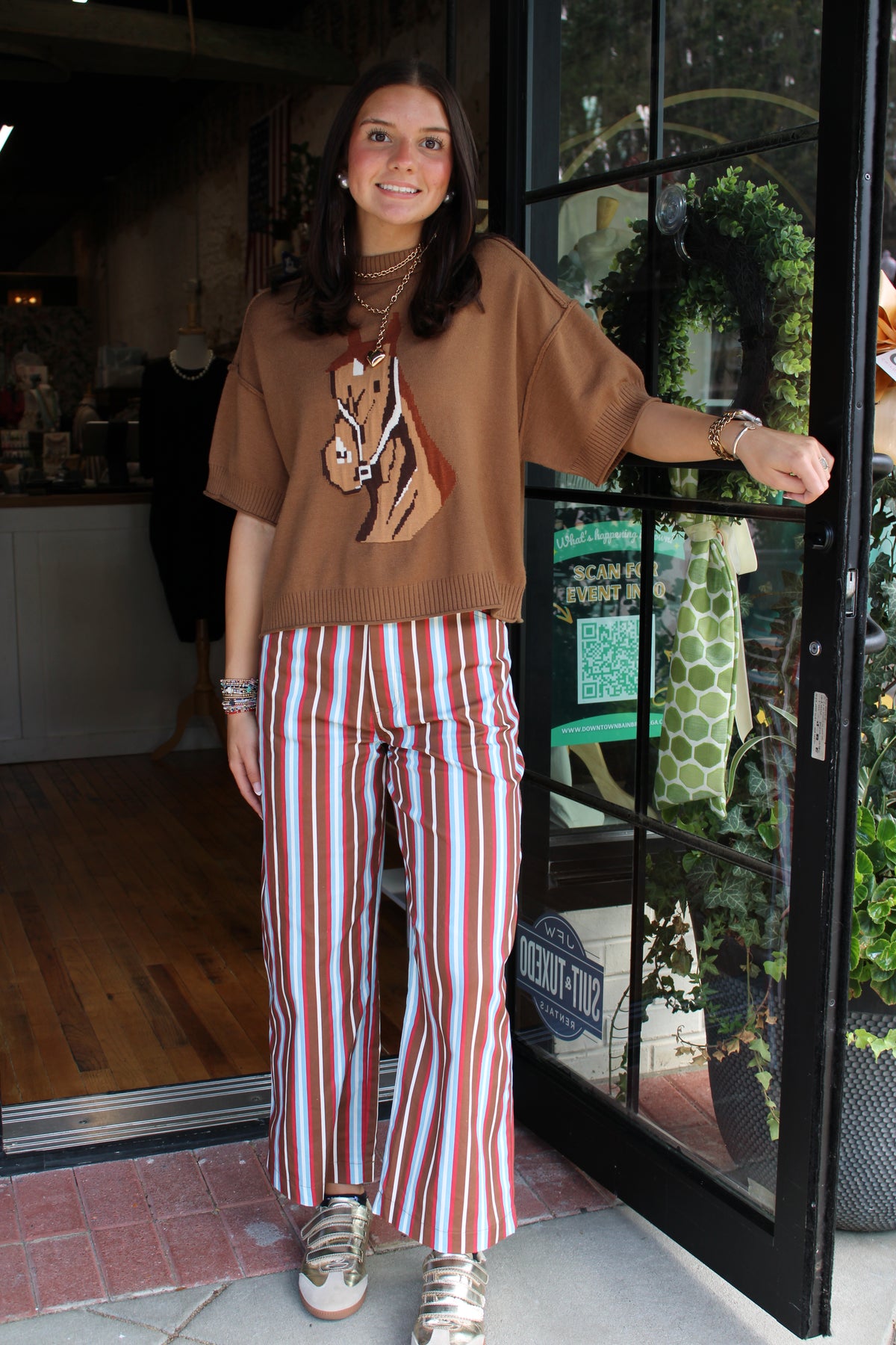 A woman stands in a storefront doorway wearing a brown cropped knit top with a horse graphic and red, white, and blue vertically striped wide-leg pants.
