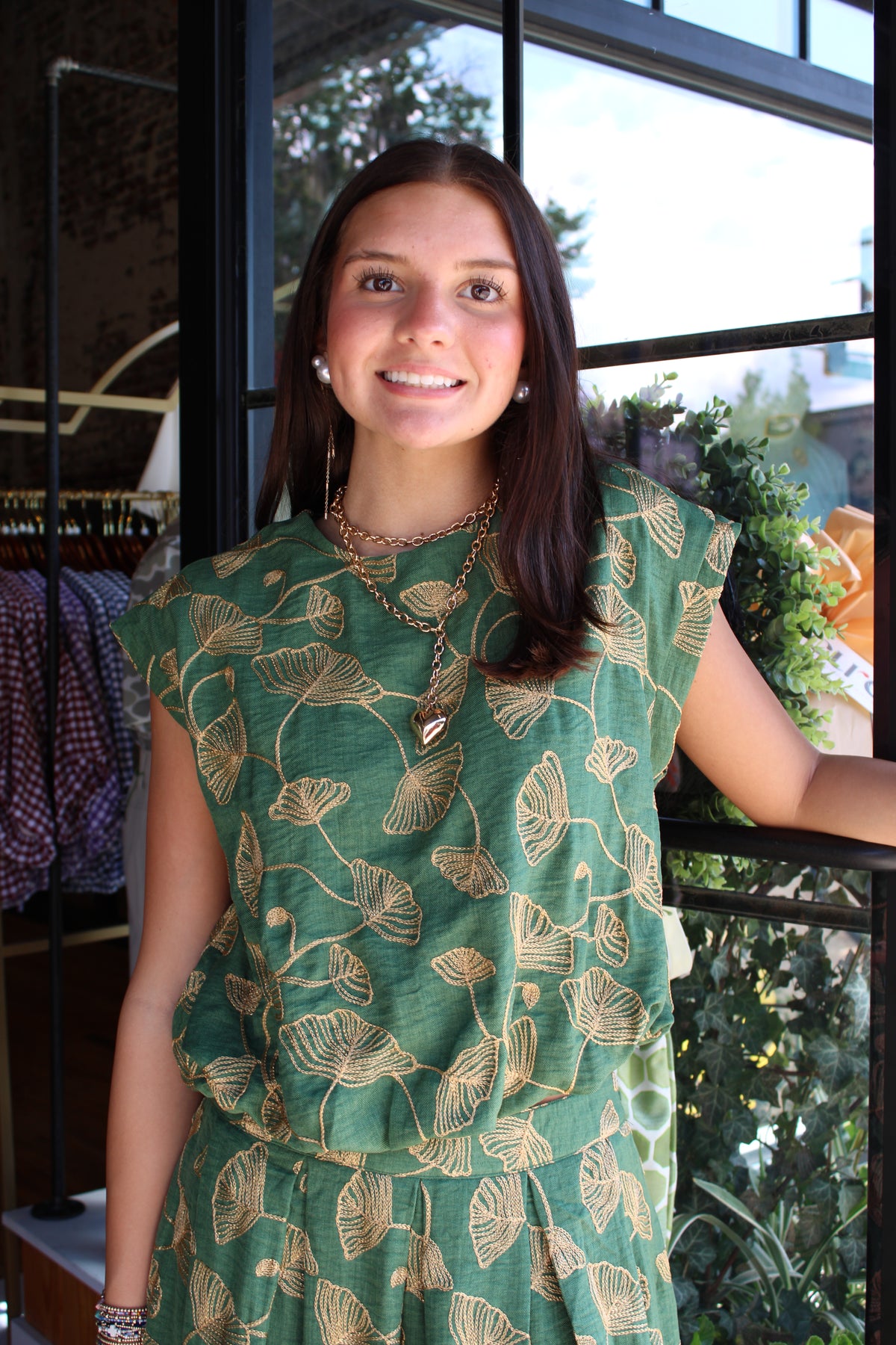 A woman wearing a green sleeveless top with a gold ginkgo leaf pattern, standing in front of a window in a boutique.