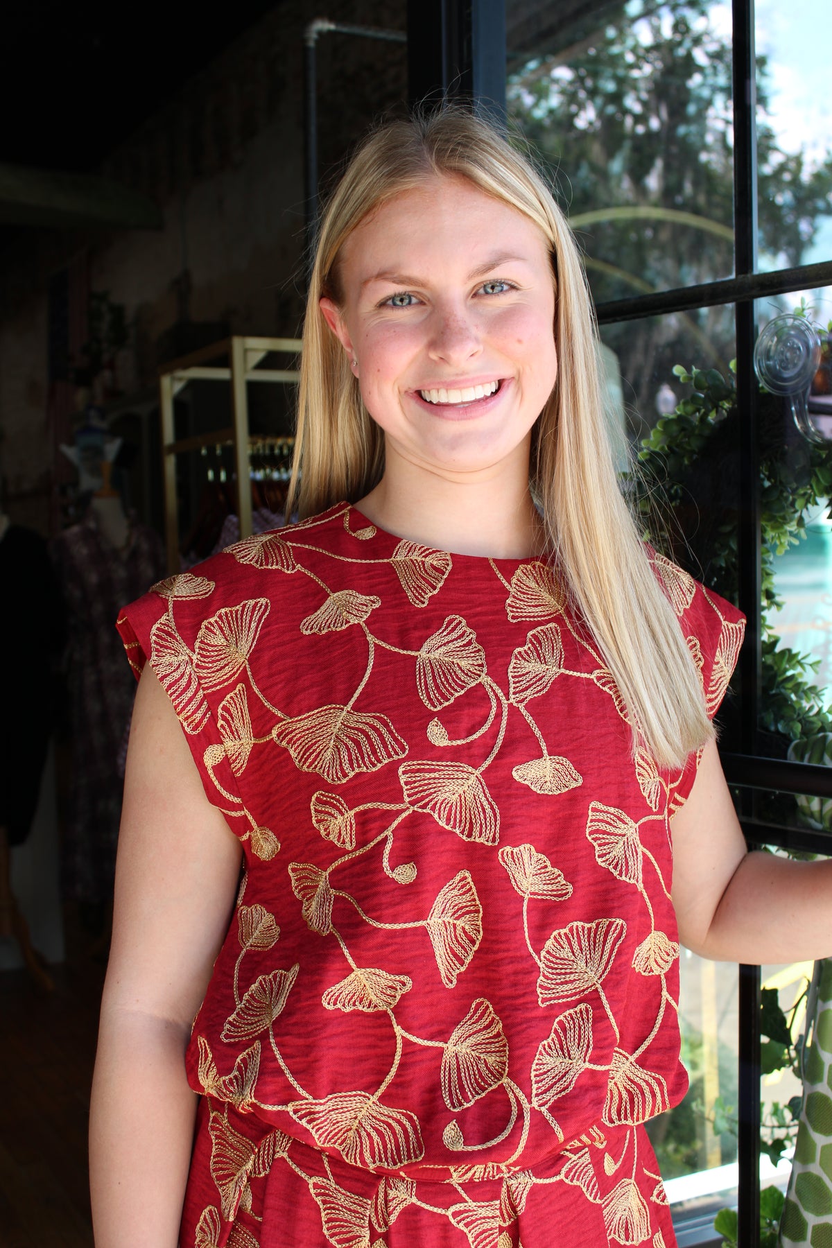 A smiling blonde woman wearing a red sleeveless top with a gold ginkgo leaf pattern, standing in front of a window.