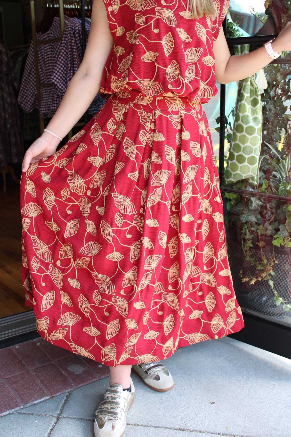 A person wearing a sleeveless red top and matching skirt with a gold leaf pattern, holding out one side of the skirt to show its drape. The person is standing on a concrete surface next to a glass door with clothing and plants visible in the background.