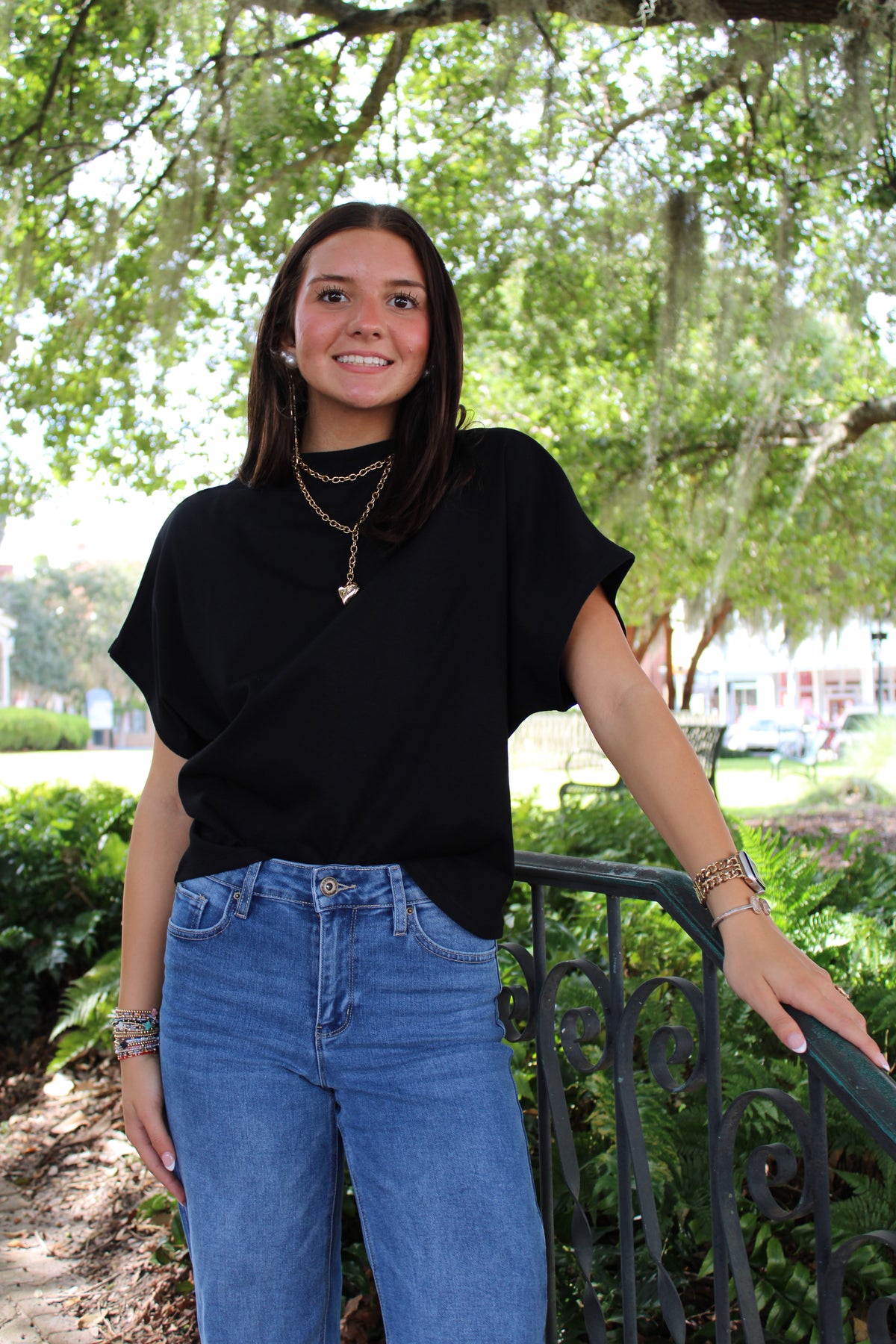 A young woman with dark hair stands outdoors in a park-like setting with trees and greenery, wearing a loose black short-sleeve top and blue jeans, with layered gold necklaces and bracelets.