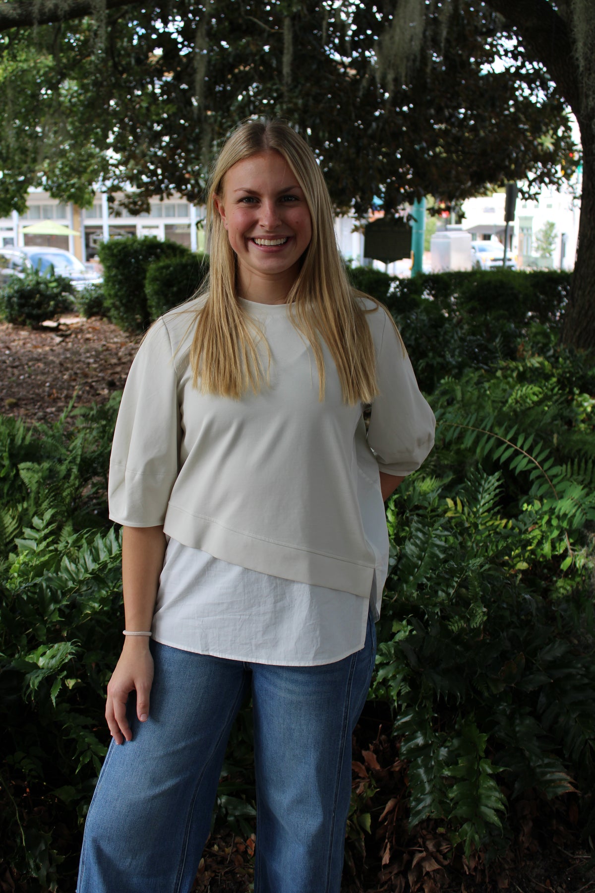 A woman with blonde hair stands outdoors in front of green bushes and trees, wearing a cream-colored asymmetrical layered top with blue jeans.