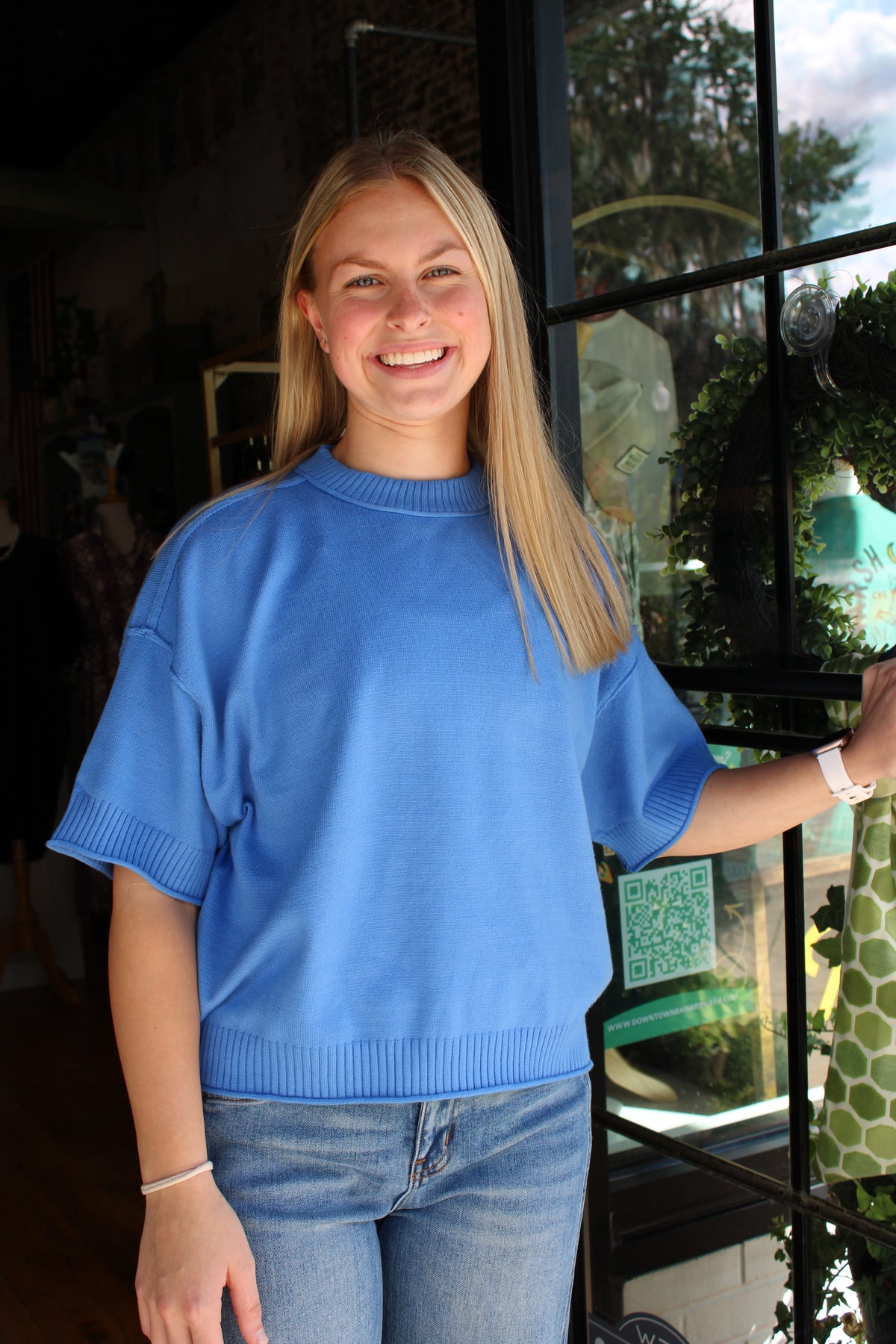 A smiling young woman with blonde hair wearing a bright blue short-sleeve crewneck sweater and light wash blue jeans, standing in front of a shop window.
