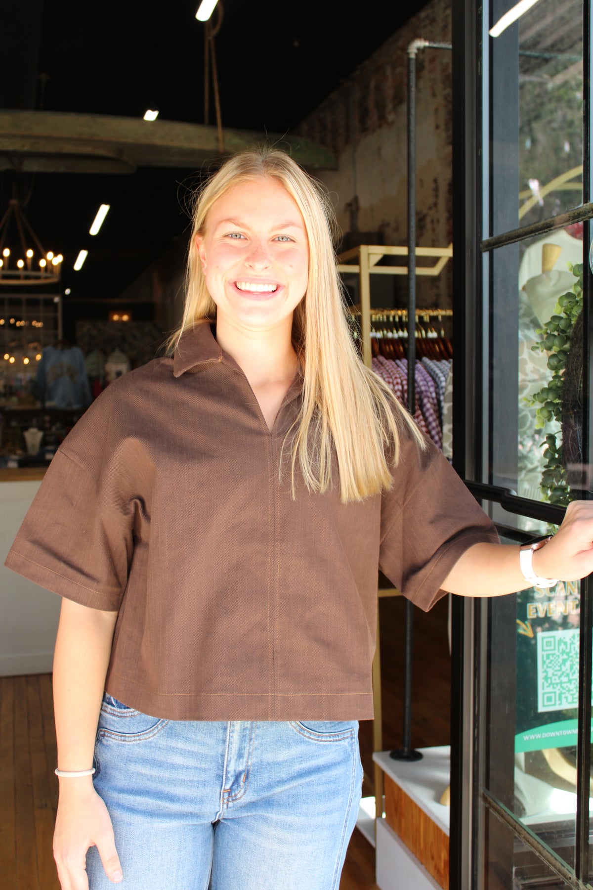 A smiling blonde woman wearing a brown short-sleeve collared button-up shirt and light blue jeans, standing in a retail store next to a glass door.