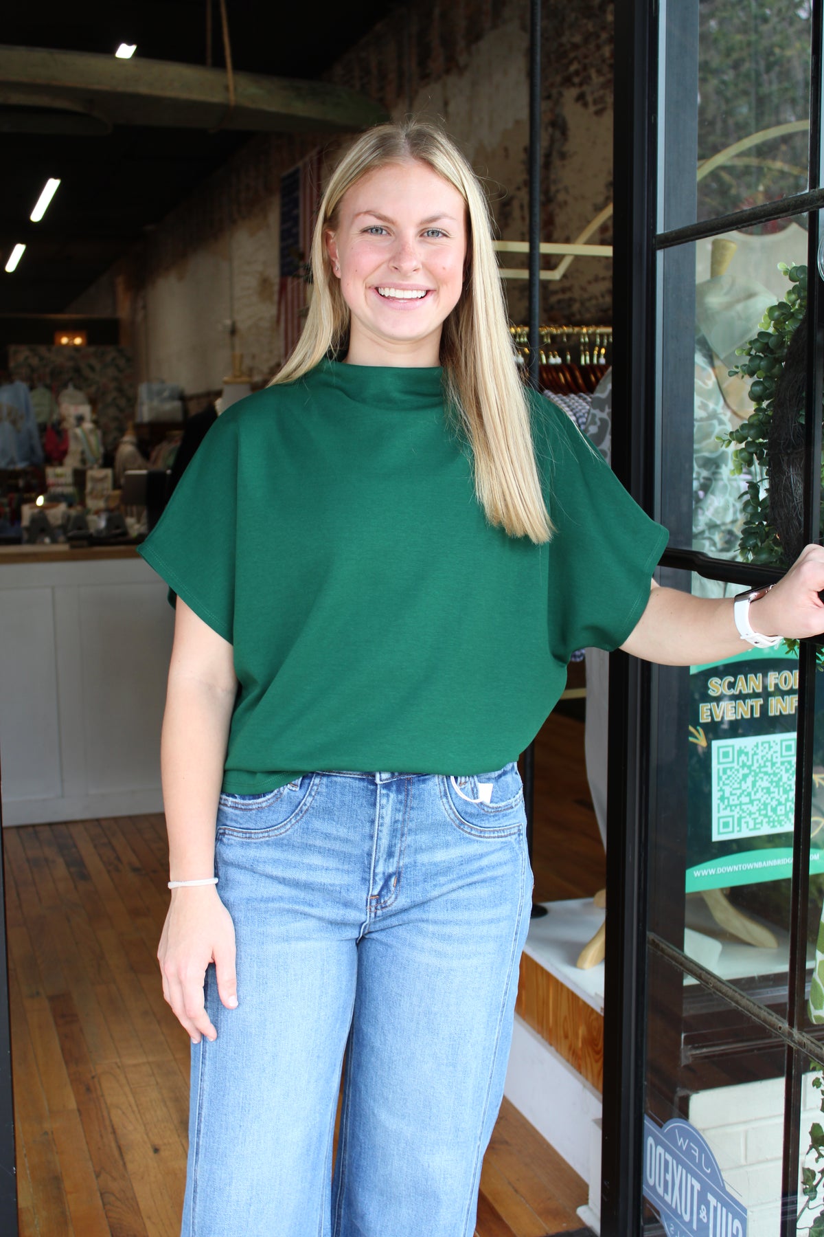A smiling blonde woman wearing a dark green short-sleeve mock neck top and light blue jeans stands in a shop doorway.