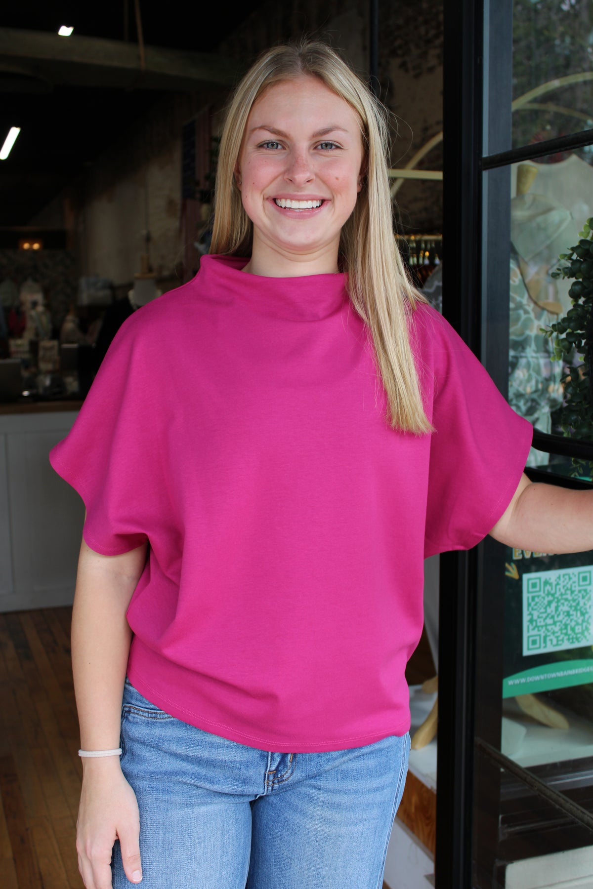 A smiling blonde woman wearing a bright pink short-sleeve mock neck top and blue jeans, standing in front of a glass door in a shop setting.