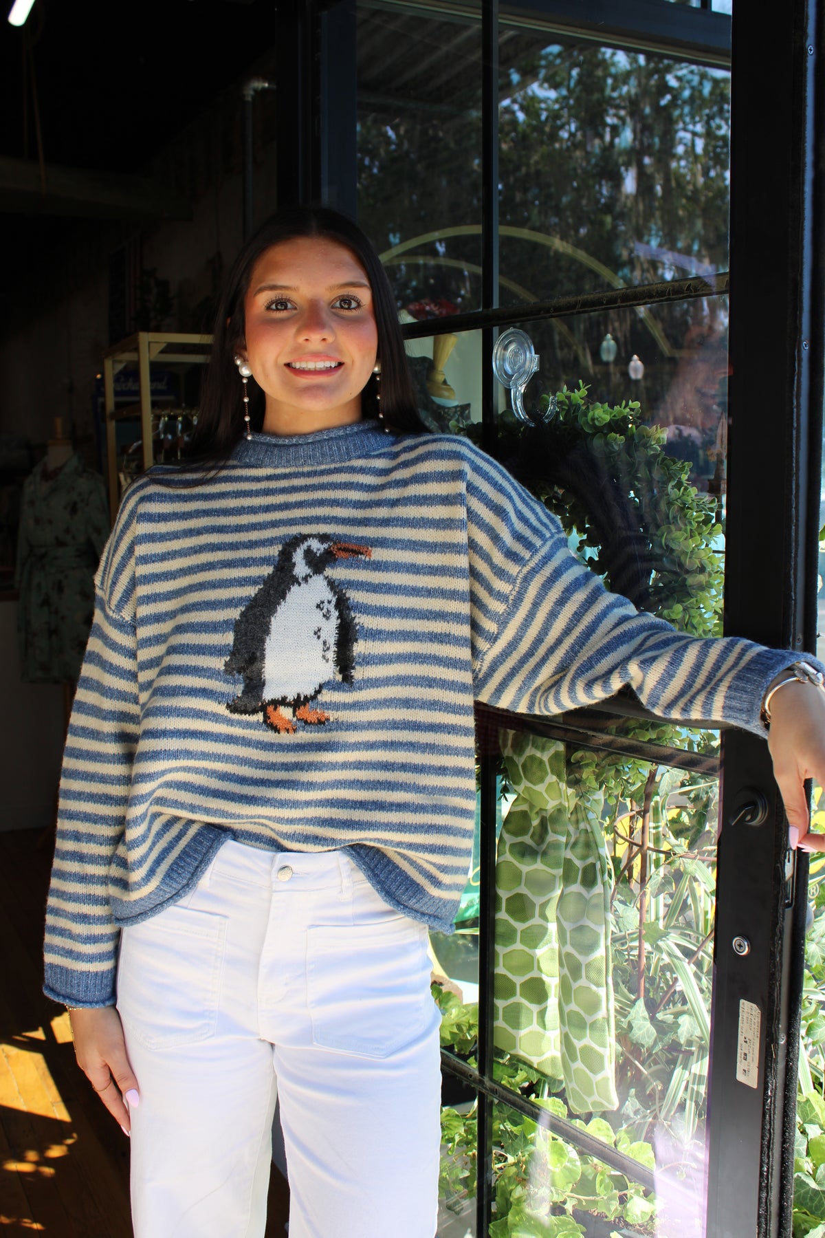 A woman wearing a blue and white striped knit sweater with a penguin graphic on the front, paired with white pants, standing in front of a glass door in a shop.