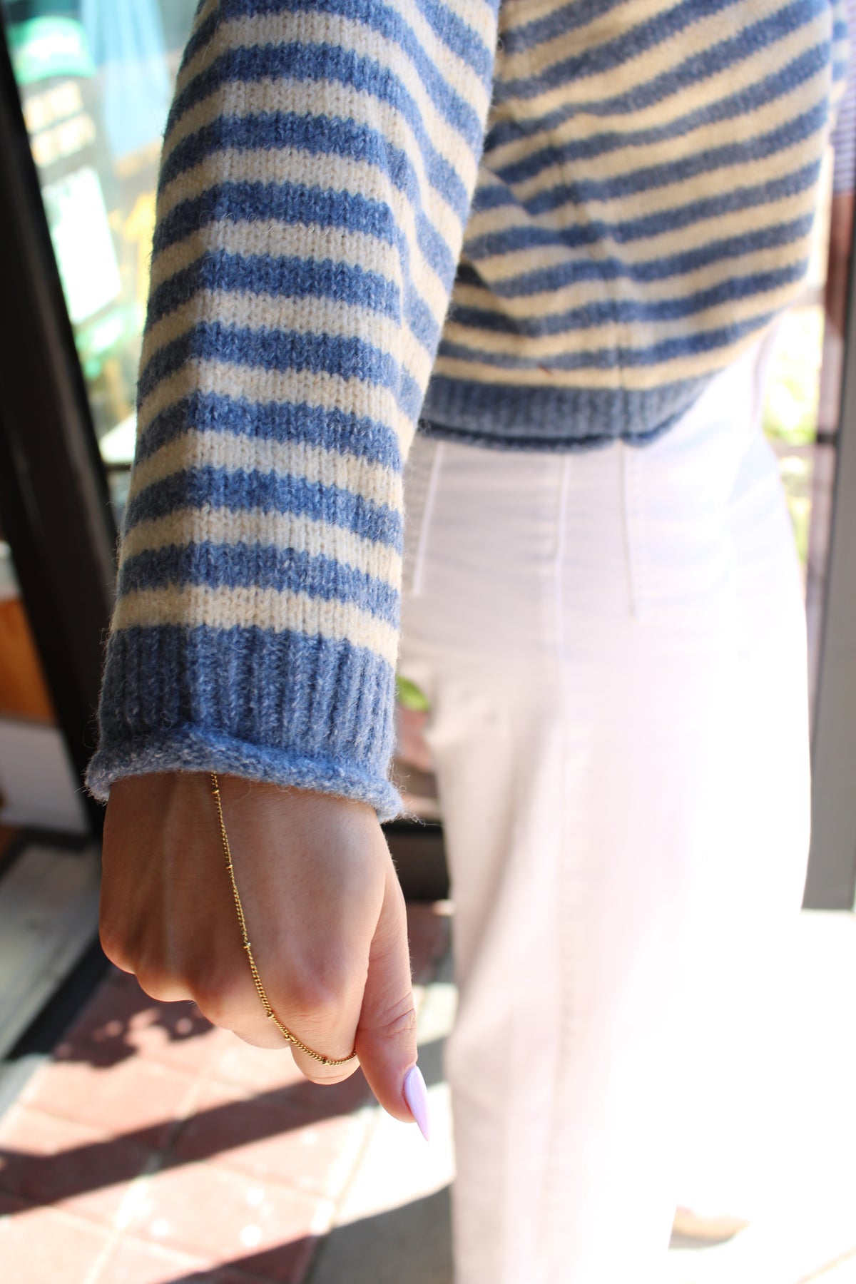 A close-up lifestyle shot of a person wearing a blue and cream striped knit sweater and white pants, with a gold chain bracelet visible on the wrist.