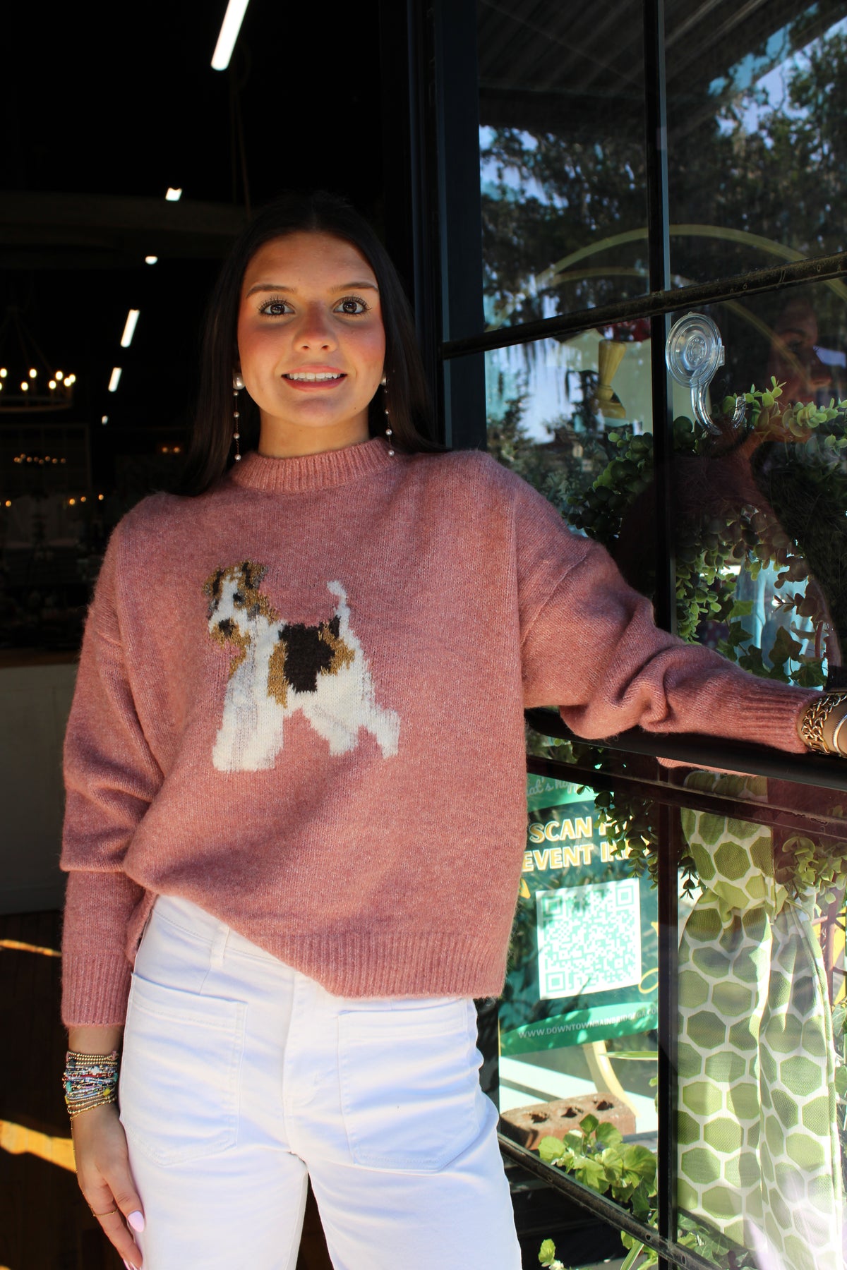 A woman wearing a dusty rose pink knit sweater with a white, brown, and black dog graphic on the front, paired with white pants, standing in front of a glass window.