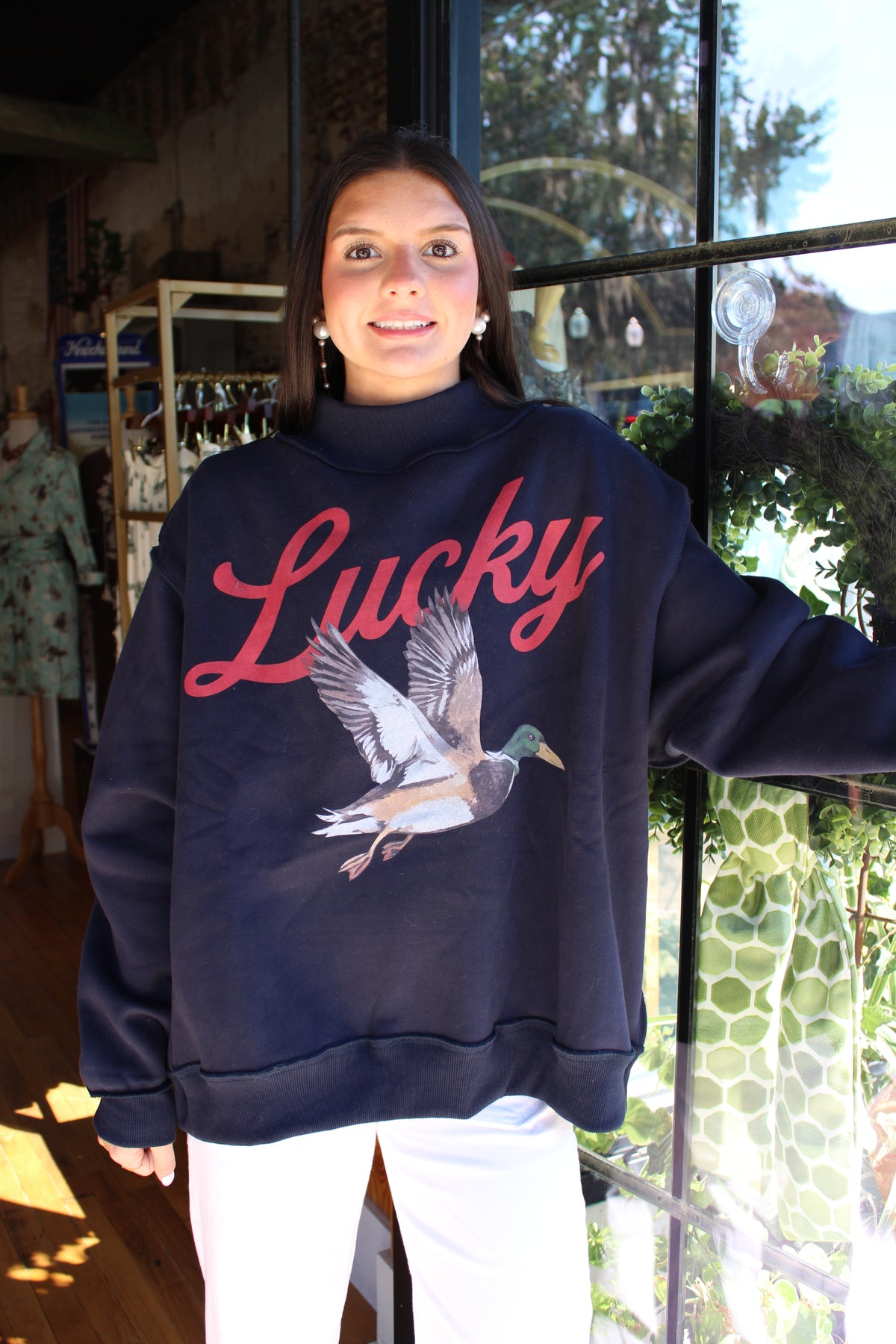 A woman wearing a navy blue sweatshirt with a red 'Lucky' script and a flying mallard duck graphic, paired with white pants, standing in front of a window.