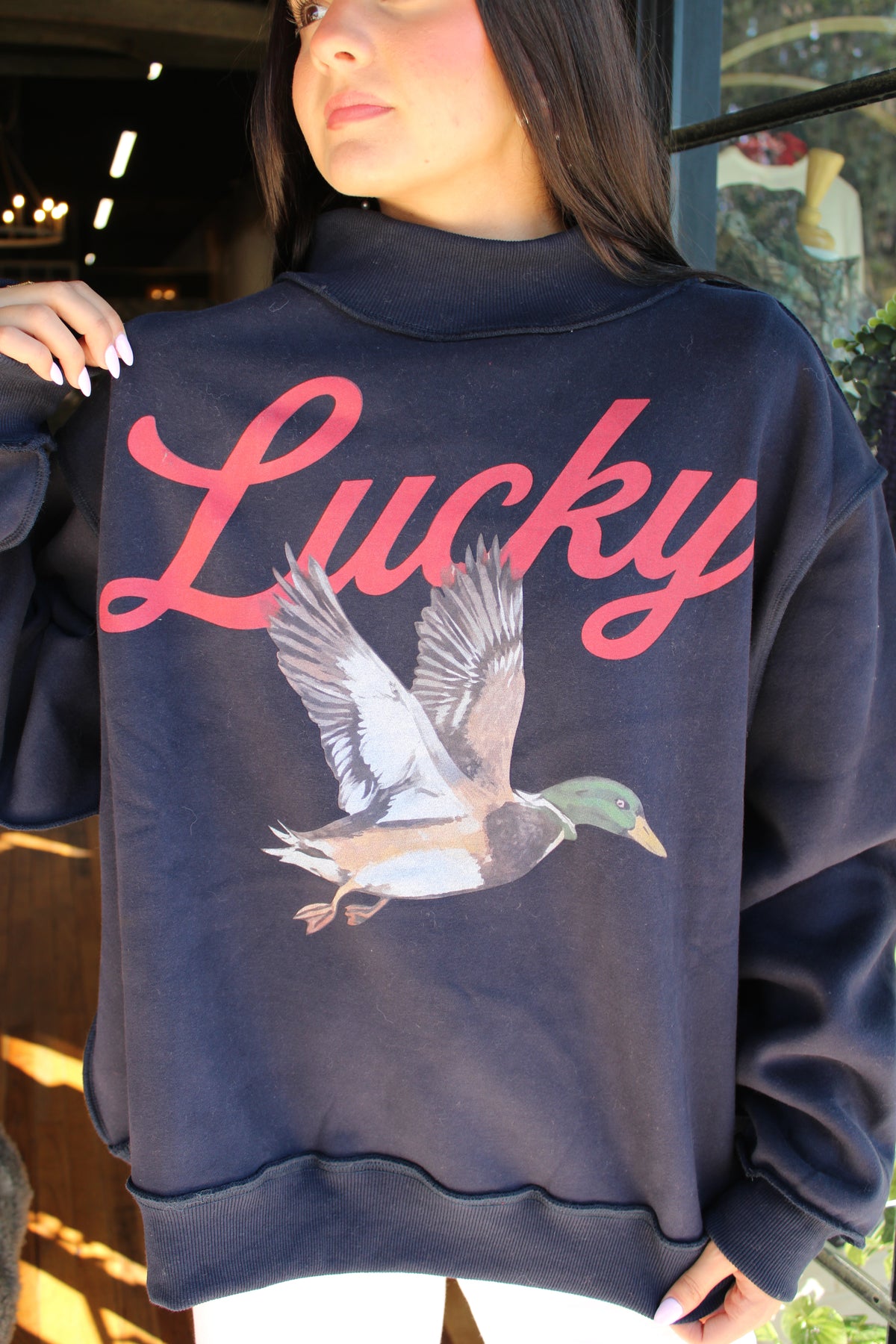 A woman wearing a navy blue sweatshirt with a large pink 'Lucky' script and a detailed mallard duck graphic on the front, photographed in a real-world setting.
