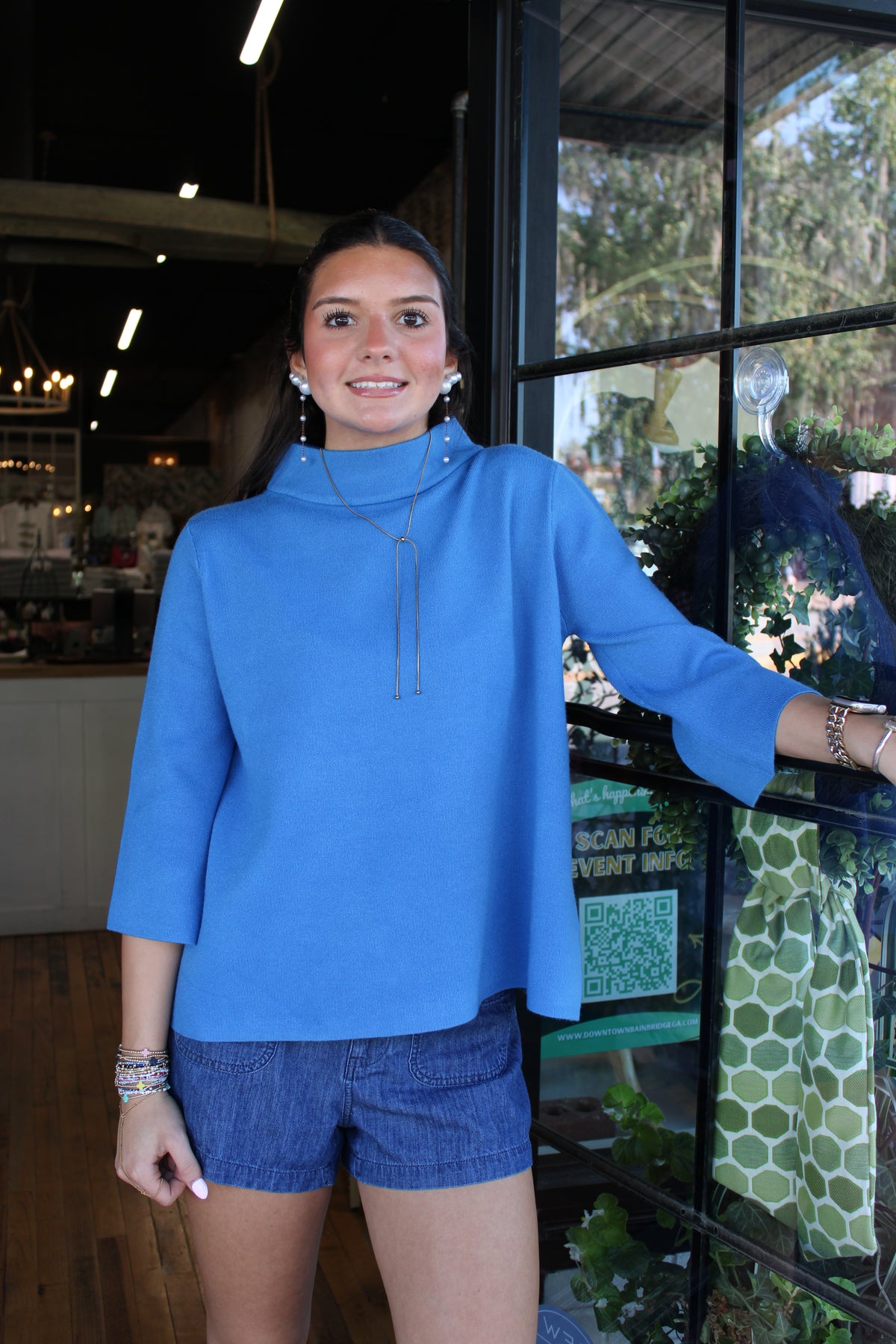 A woman wearing a bright blue mock-neck long-sleeve top and denim shorts, standing in front of a glass window display inside a boutique.