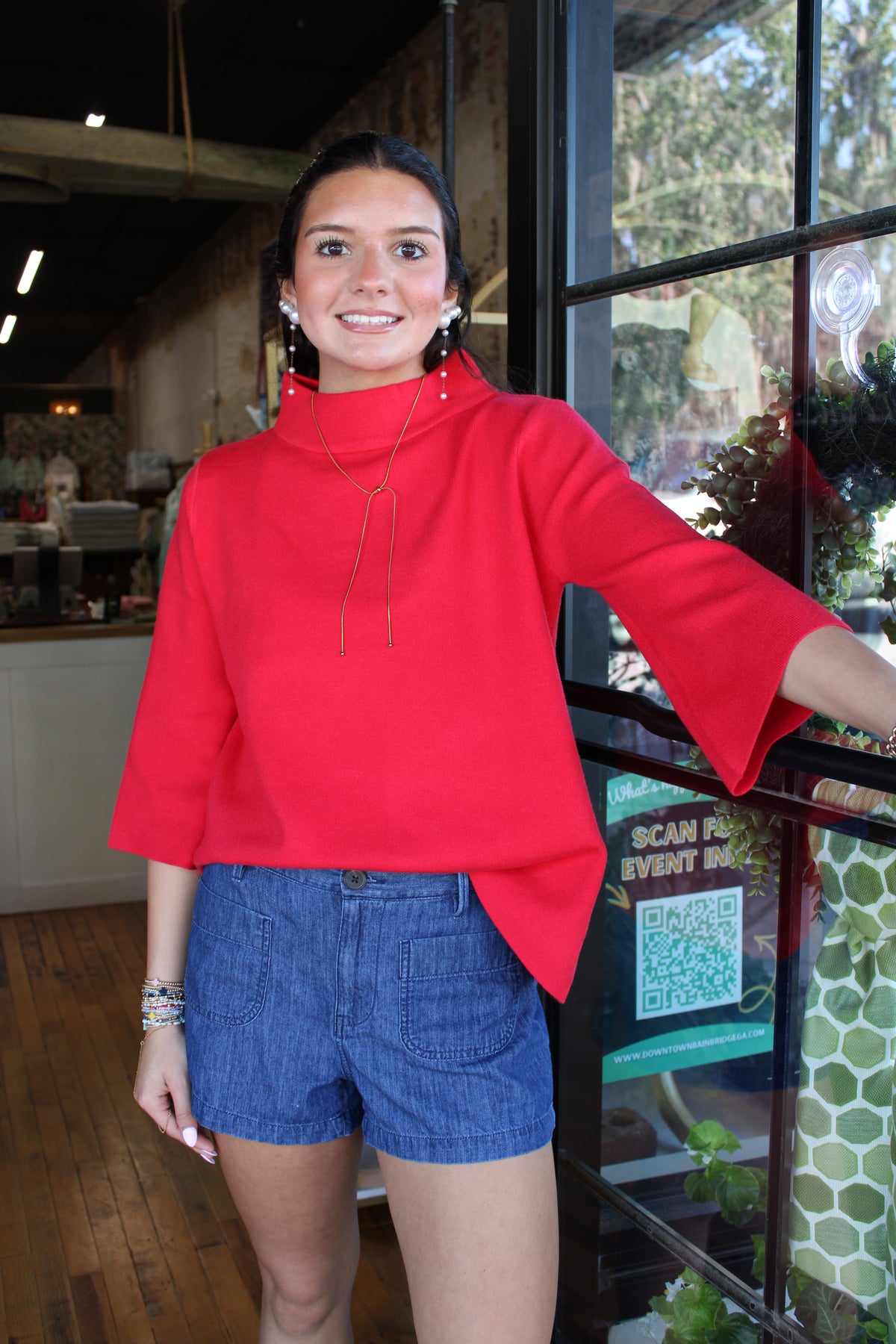 A woman wearing a bright red mock-neck top with three-quarter bell sleeves and blue denim shorts, standing in a cafe or boutique next to a window.