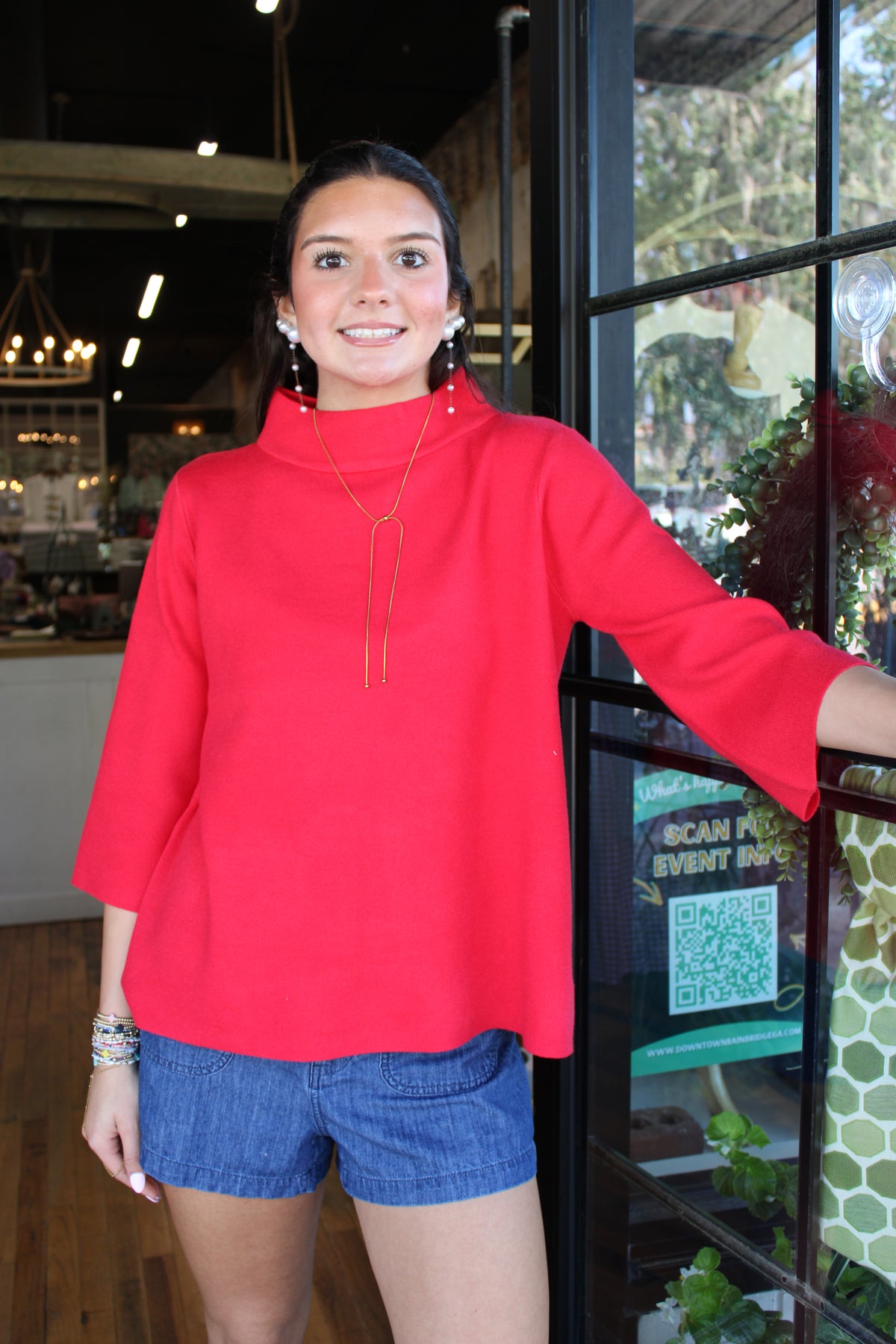 A woman wearing a bright red mock-neck top with three-quarter bell sleeves and blue denim shorts, standing in front of a glass window in a shop.
