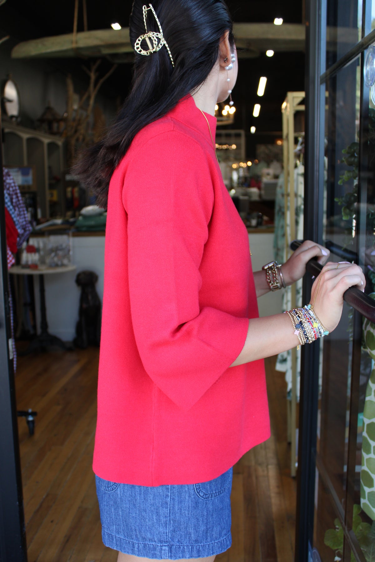A woman wearing a bright red mock-neck top and a denim short, standing in a shop doorway.
