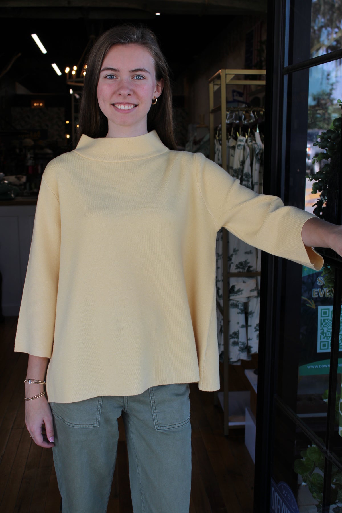 A woman wearing a pale yellow mock-neck long-sleeve top and olive green pants, standing in a shop next to a glass door.