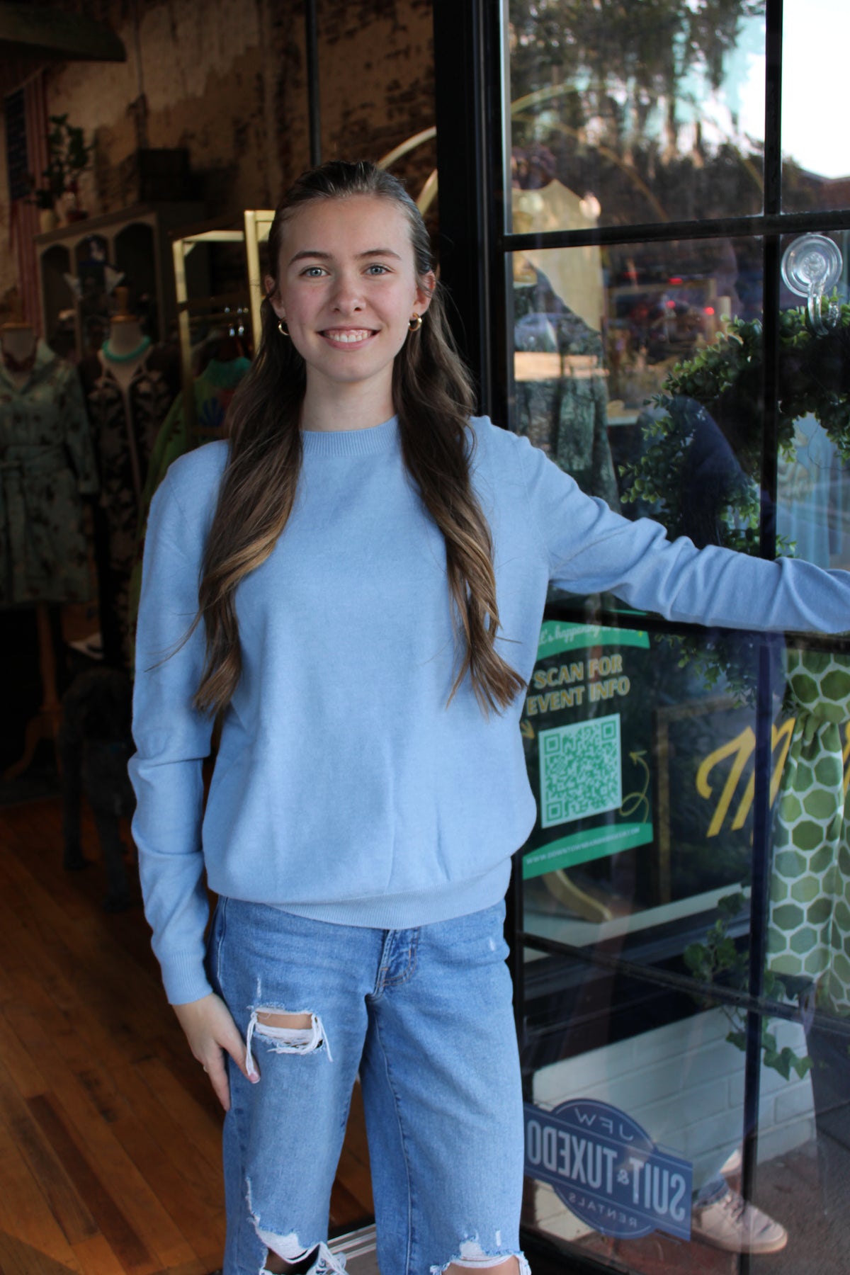 A young woman with long brown hair stands in a clothing store doorway wearing a light blue crewneck sweatshirt and distressed light blue jeans.