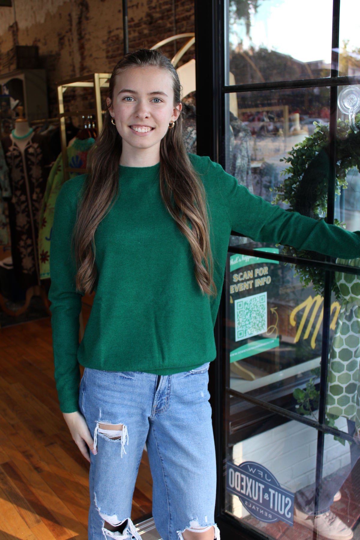 A young woman with long brown hair stands in a clothing store doorway wearing a bright green long-sleeve mock-neck sweater and light-wash ripped blue jeans.