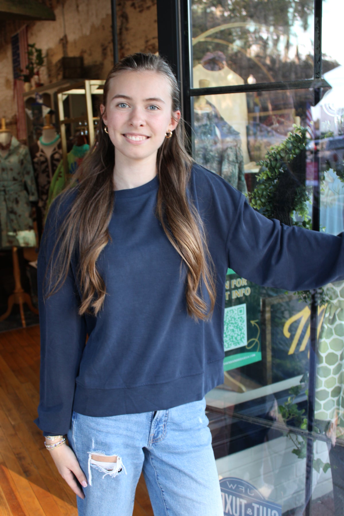 A young woman with long brown hair stands in a shop wearing a navy blue crewneck sweatshirt and light-wash ripped jeans, smiling and gesturing with her right arm toward a window display.