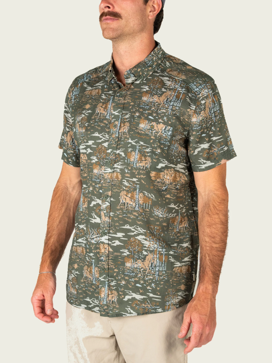 A man wearing a short-sleeve button-up shirt with an allover deer hunting camouflage pattern in olive green, brown, and beige, paired with khaki pants, against a light off-white background.