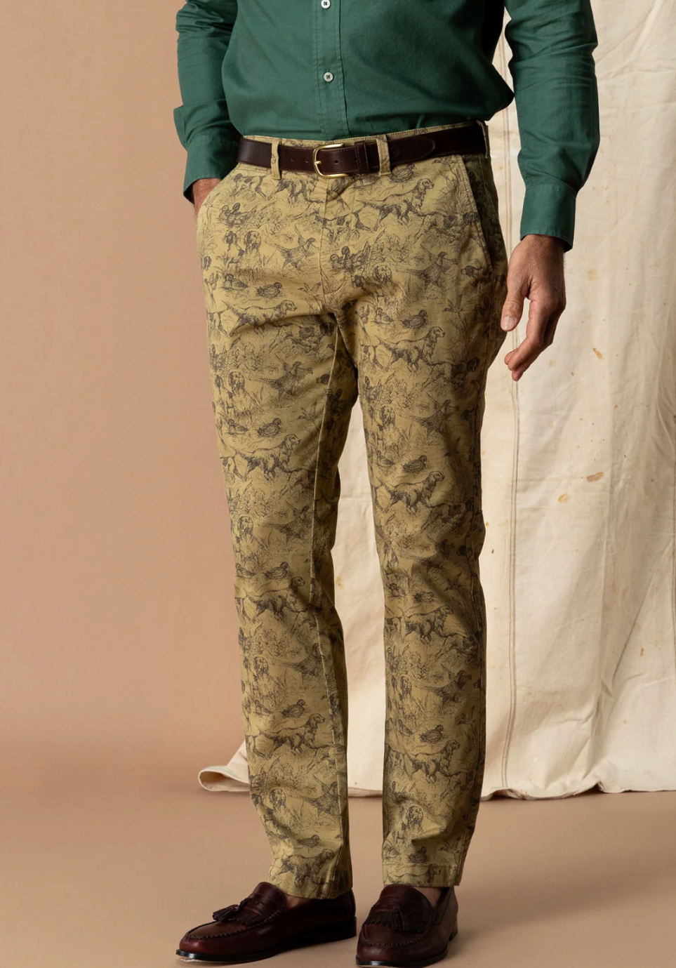A man wearing slim-fit trousers with an abstract dark brown and olive-green pattern on a beige base, styled with a green shirt, brown belt, and dark brown loafers.