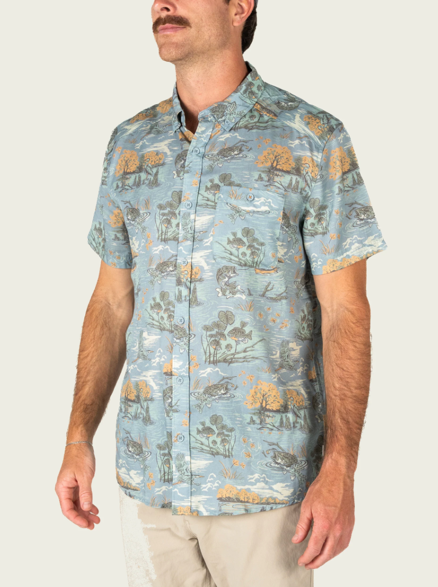 A man wearing a light blue short-sleeve button-up shirt with a detailed landscape & fish print in muted green, brown, and orange tones, paired with beige pants against a solid light background.