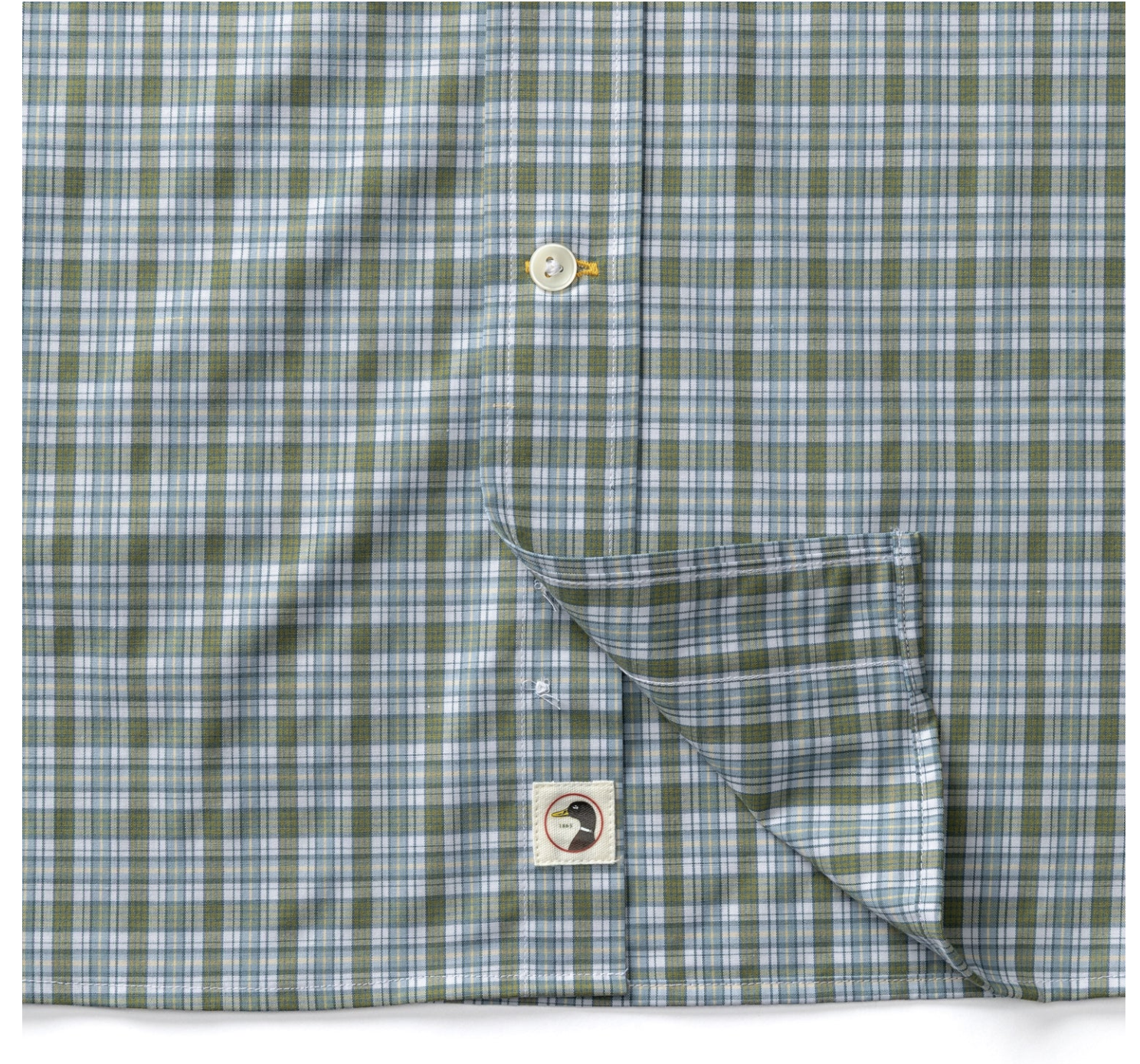 A close-up of a plaid button-down shirt in sage green, light blue, and white, with the cuff turned up to show the inner lining and a small brand patch.