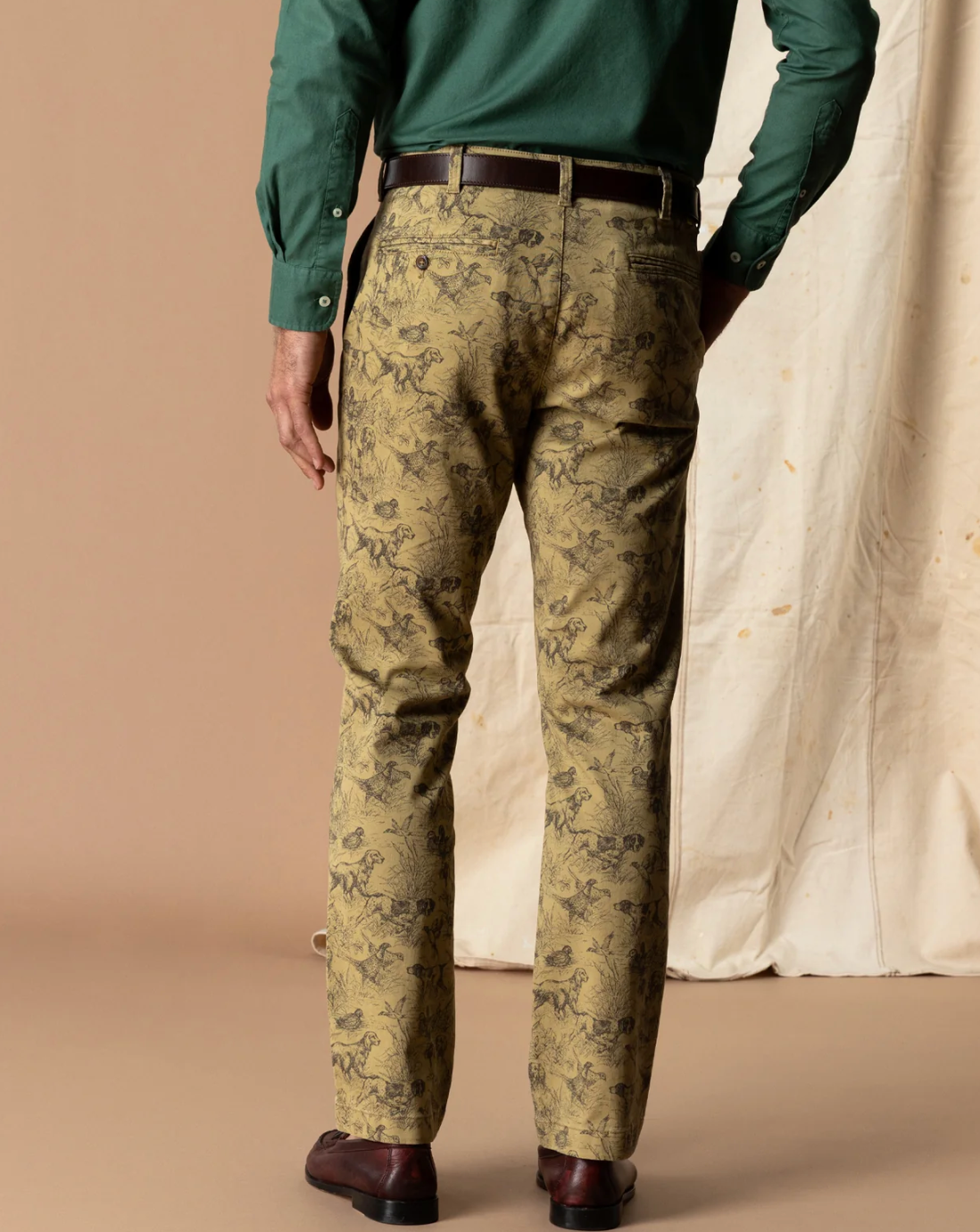 A man is shown from the back wearing slim-fit trousers with an all-over toile-style floral print in olive green and beige, paired with a dark brown belt and dark brown shoes.