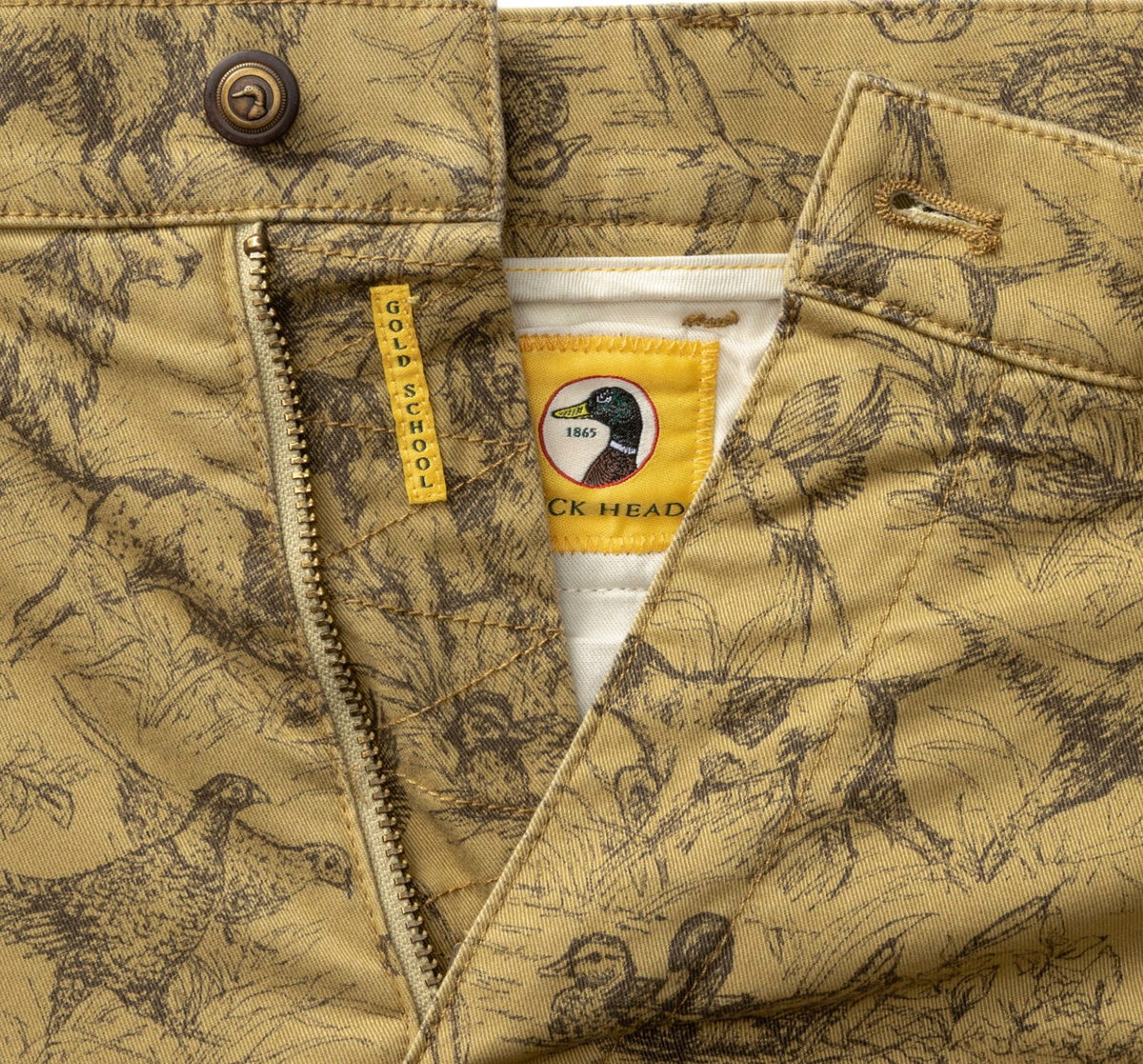 Close-up of a tan and brown duck hunting patterned jacket with a zipper, button, and a yellow neck label featuring a duck head logo and the text 'Duck Head'.