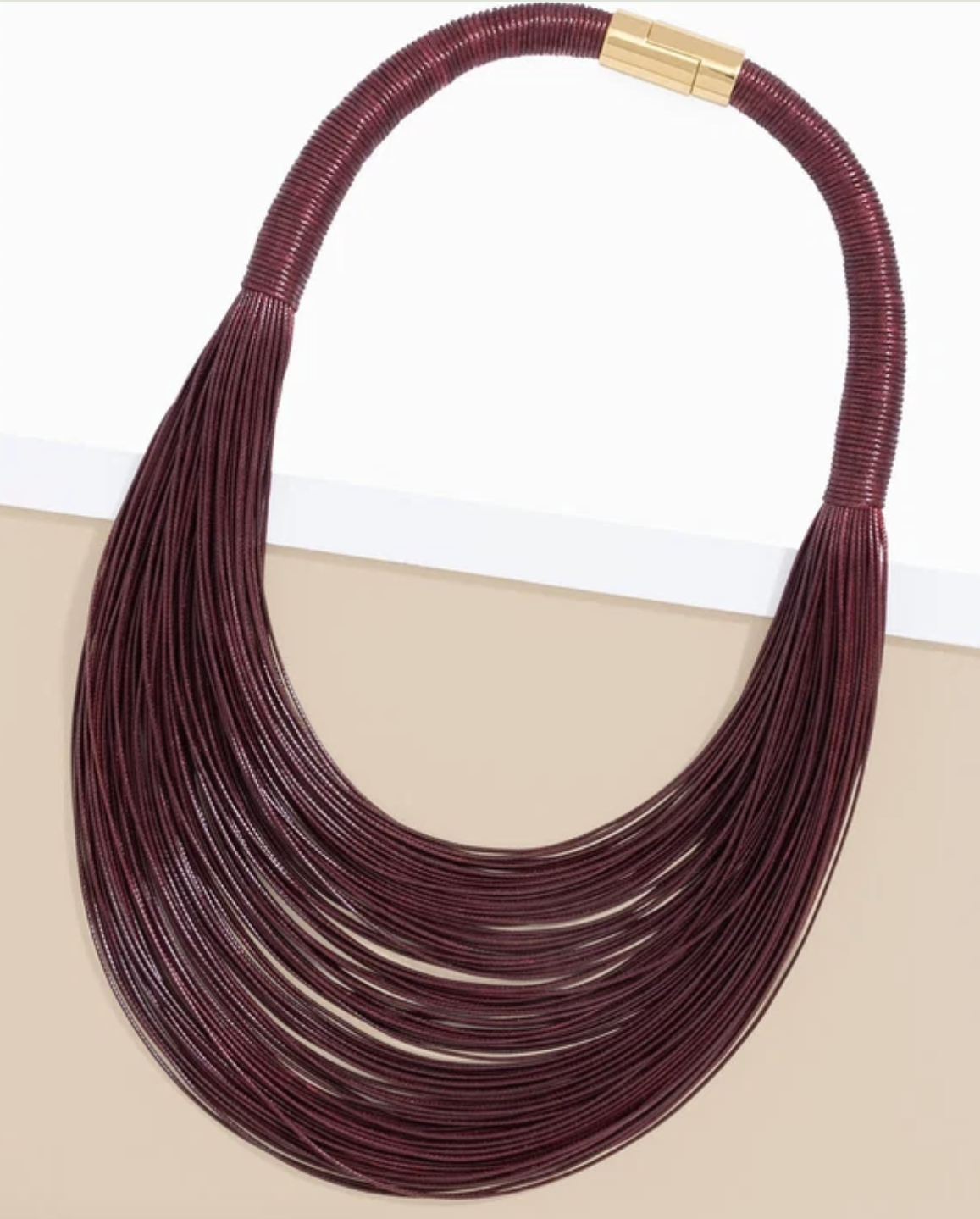 A multi-strand statement necklace made of dark burgundy cord with a gold clasp, displayed on a split white and beige background.