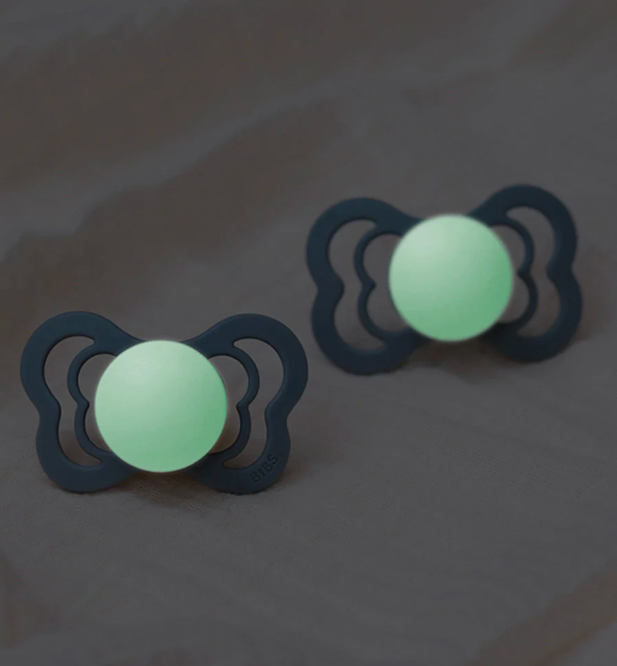 Two glow-in-the-dark pacifiers with dark gray silencers and luminous mint-green centers, displayed on a textured gray surface.