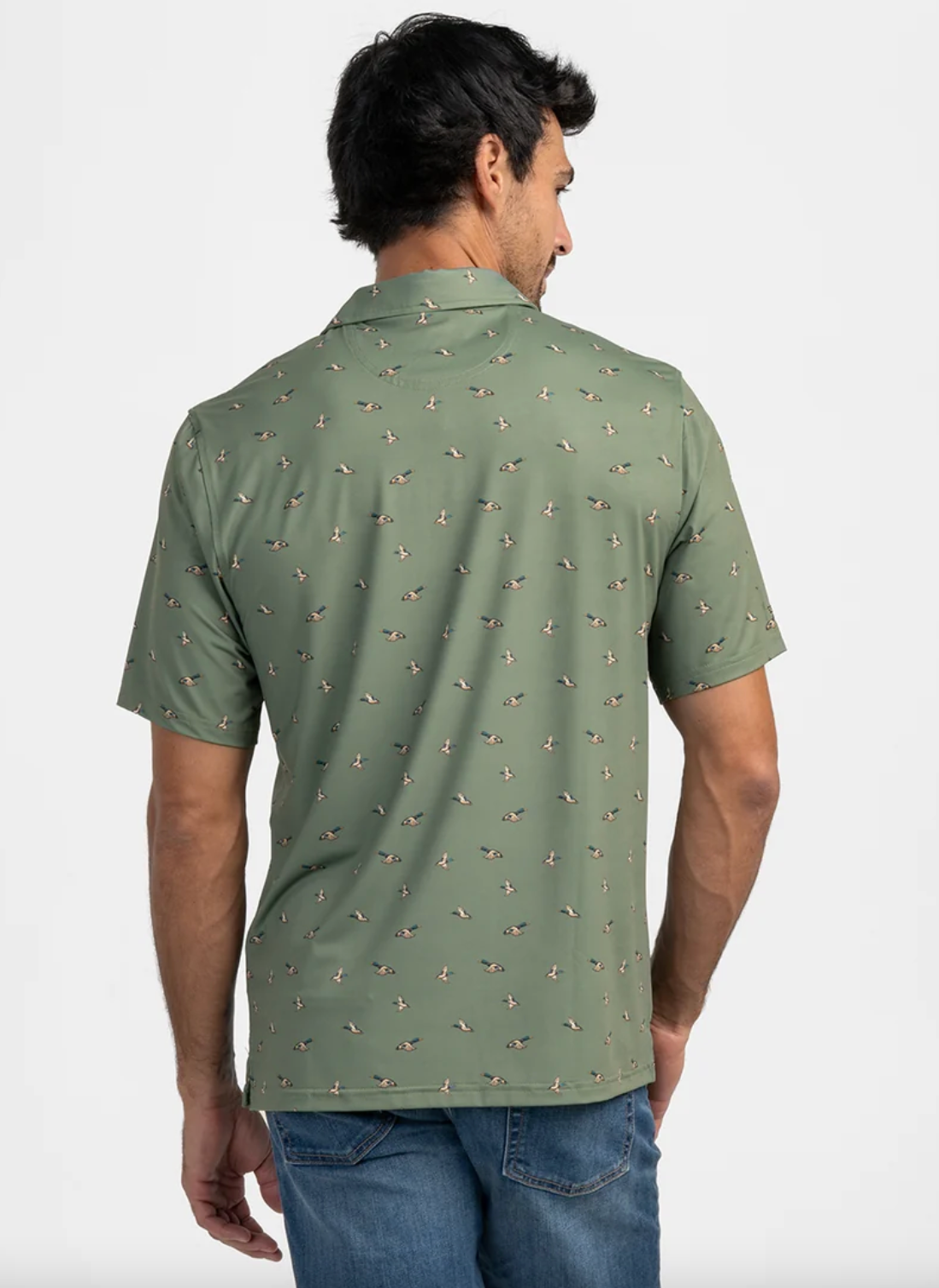 A man viewed from the back wearing a short-sleeve polo shirt in sage green with a small all-over mallard print, paired with blue jeans against a light gray studio background.