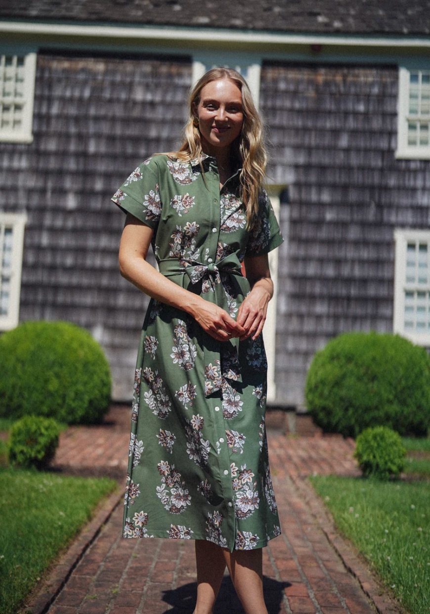 A woman stands on a brick path in front of a shingled house, wearing a green short-sleeve midi dress with a white and browns floral print and a tie waist.