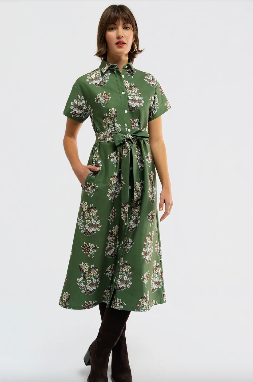 A woman modeling a green short-sleeve midi shirt dress with a white and beige floral print, a button-down front, and a matching waist tie, paired with dark brown knee-high boots against a light gray studio background.