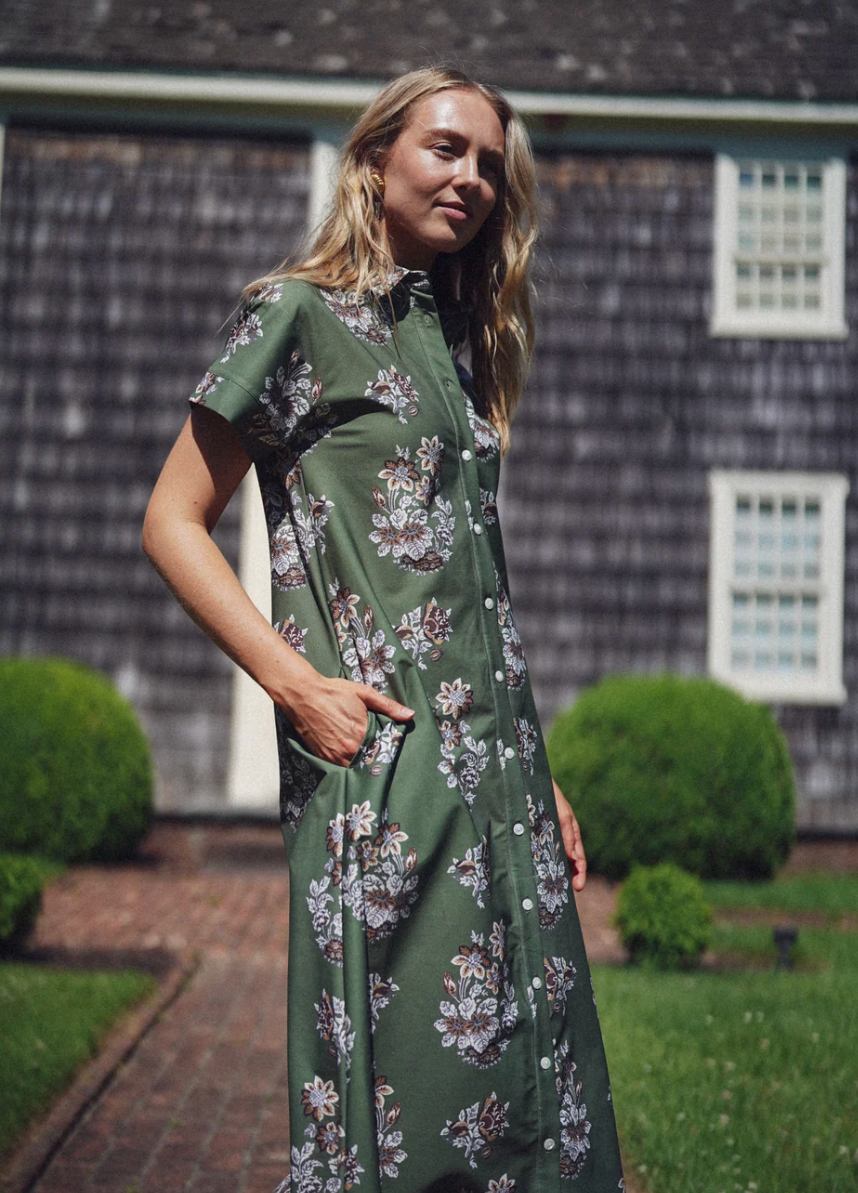 A woman wearing a green short-sleeve button-front dress with a white and brown floral pattern, standing outdoors in front of a dark shingled house with manicured green bushes.