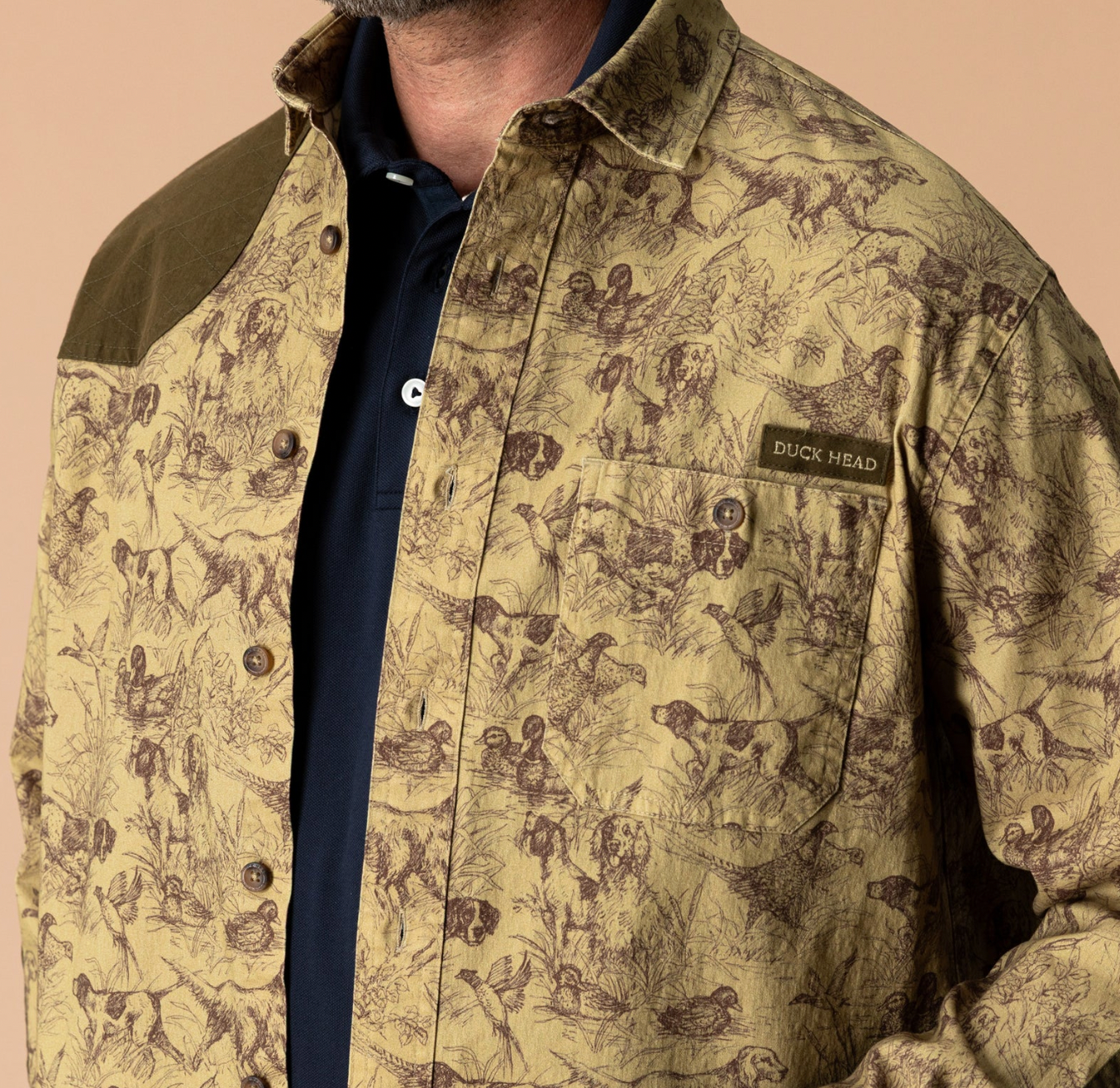 A close-up of a man wearing a Duck Head button-up shirt with a detailed dog hunting sketch pattern in brown on a tan background, featuring a solid brown collar and shoulder yoke, layered over a navy blue polo shirt.